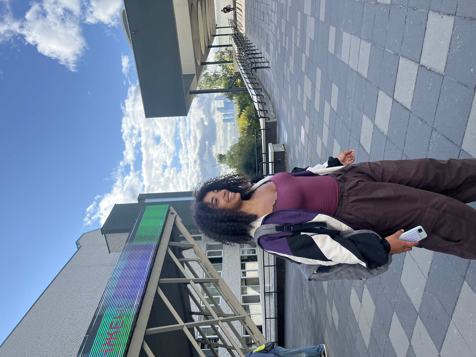 A young woman with curly dark hair and a light complexion is walking in an outdoor urban environment. She is wearing a burgundy tank top, a purple, black, and white jacket, and brown pants. She has a gray backpack and is holding a light blue smartphone in her right hand. The ground is paved with large square tiles in a checkerboard pattern. In the background, there is a modern building with a large LED screen displaying green and purple lights, a metal staircase, and trees. In the far distance, the Moscow skyline with skyscrapers is visible. The sky is blue with white clouds.