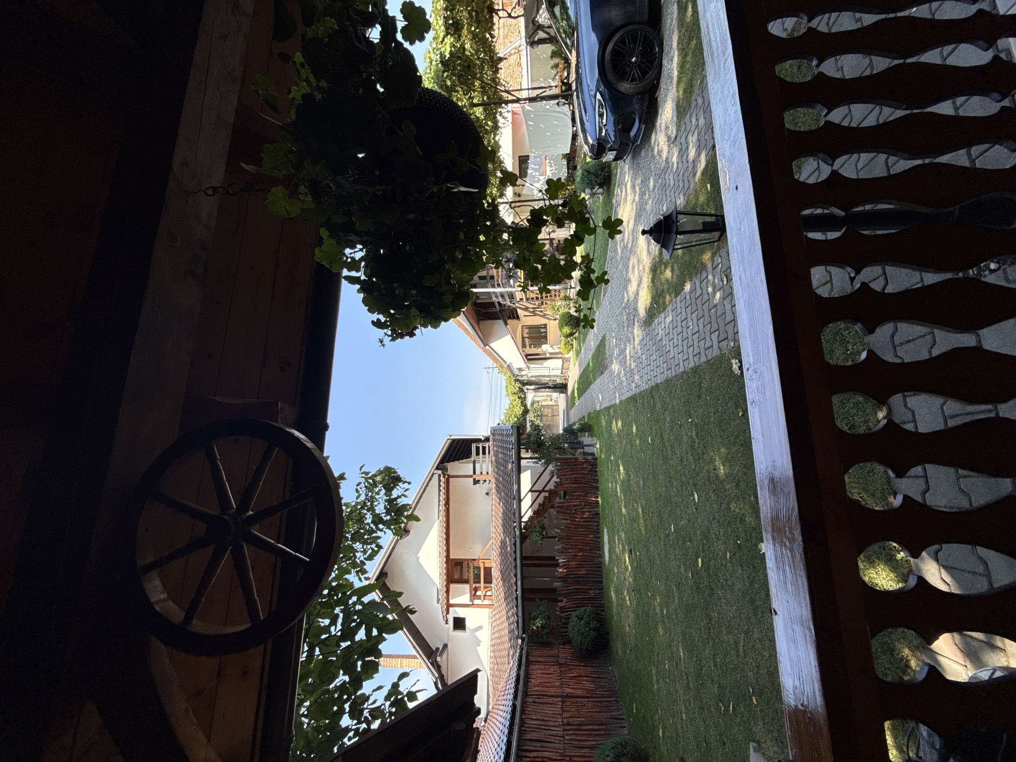 The image shows a view from a wooden porch overlooking a residential property in Cârțișoara, Romania. In the foreground, the wooden railing of the porch is visible, with decorative cutouts filled with cobblestone and grass. Above, a wagon wheel is mounted on the wooden ceiling of the porch. To the right, a hanging basket filled with green plants is suspended from the porch's roof.

The property features a well-maintained lawn bordered by a rustic wooden fence. A paved walkway, made of interlocking bricks, leads away from the porch and past a modern, dark blue sedan parked on the right side of the walkway. A black lantern sits beside the walkway.

Two houses are visible. The house on the left is a white, two-story building with a prominent chimney and a wooden balcony on the upper level. The house on the right appears to be a single-story dwelling with a white facade. Trees and foliage are scattered throughout the property, and the sky is clear and blue, indicating a sunny day. The overall impression is of a peaceful, residential setting with a blend of rustic and modern elements.