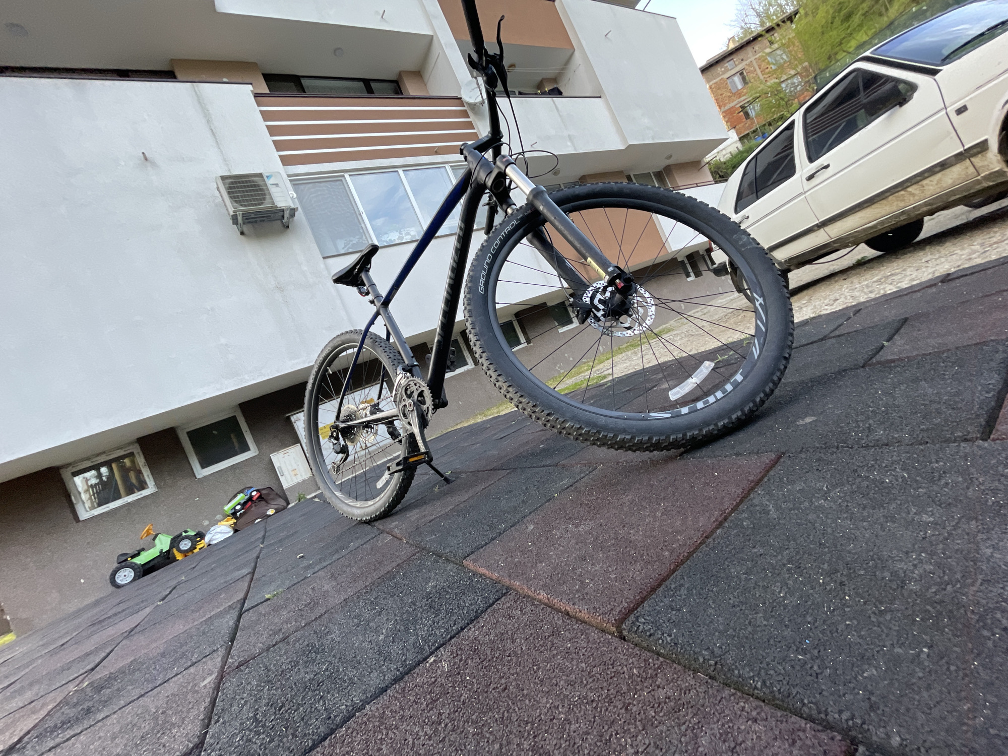 A bicycle is parked on an outdoor rubber tile surface near a building. Other buildings and a parked white Volkswagen Golf are in the background. The building has a white facade with brown horizontal accents. A window air conditioner is mounted on the building's exterior wall. A few children's toys are visible near the building on the left. The location is Zlatograd, Bulgaria. There are no people visible in the image.