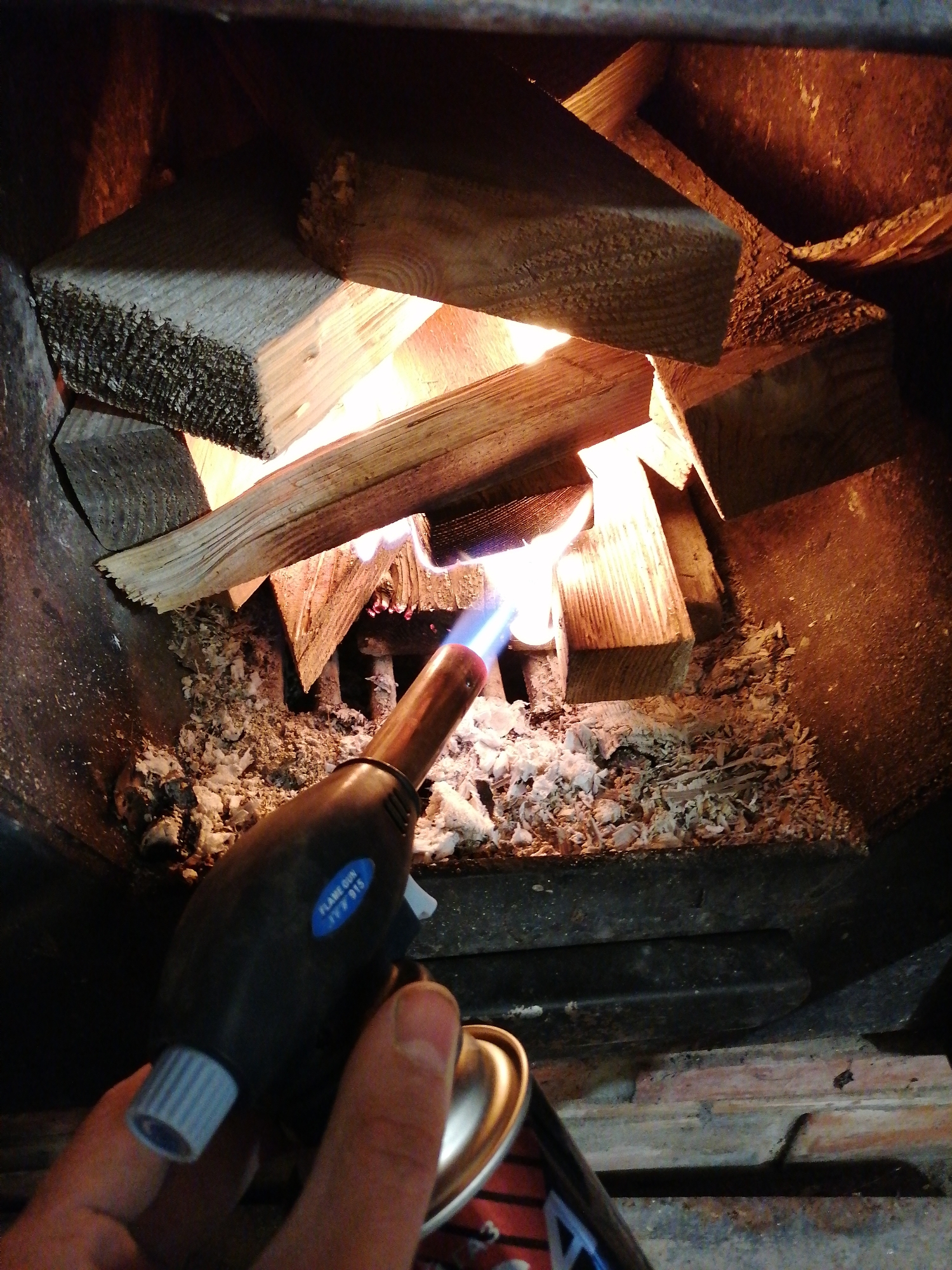 A hand holds a lit blowtorch with a blue flame, aimed at wood kindling inside a dark, enclosed space, likely a fireplace or wood-burning stove. The interior is lined with dark metal and filled with wood shavings. The wood is stacked and partially lit by flames. The environment suggests a residential or rural setting.