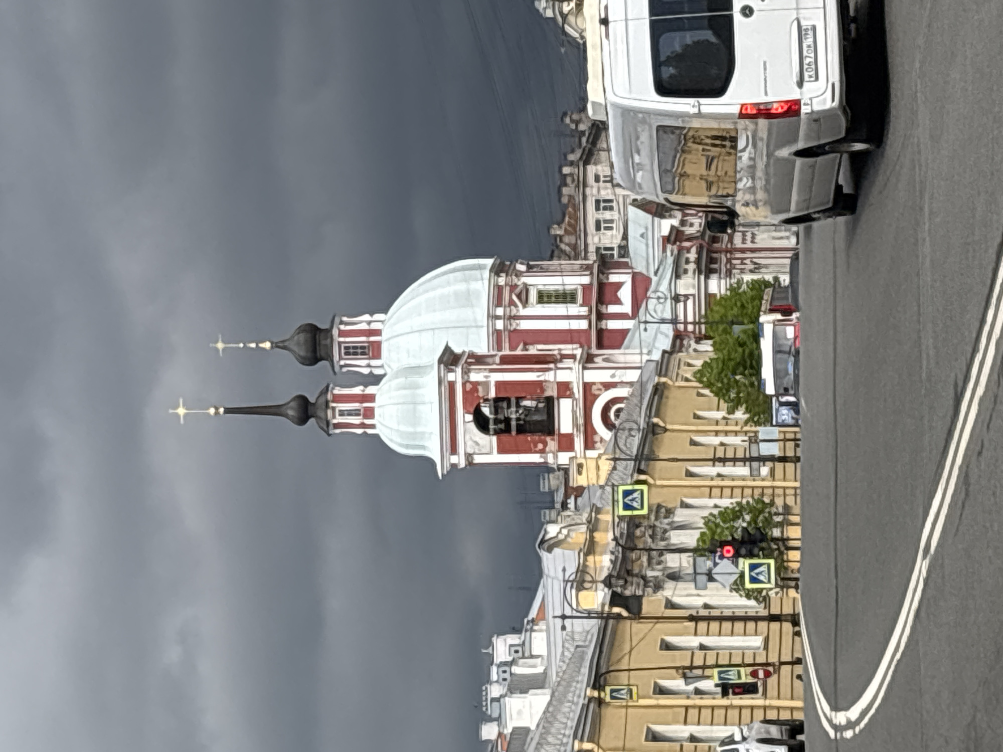 The image depicts the Church of Saints Simeon and Anna in Saint Petersburg, Russia, under a cloudy, dark sky. A white van occupies the upper right of the frame, obscuring some of the building's details. The church features white domes and red and white facades, with crosses atop its spires. Buildings line the street adjacent to the church, showing a mix of architectural styles. Street signs and traffic signals are also visible along the street. The angle suggests the photo was taken from a moving vehicle on the road.