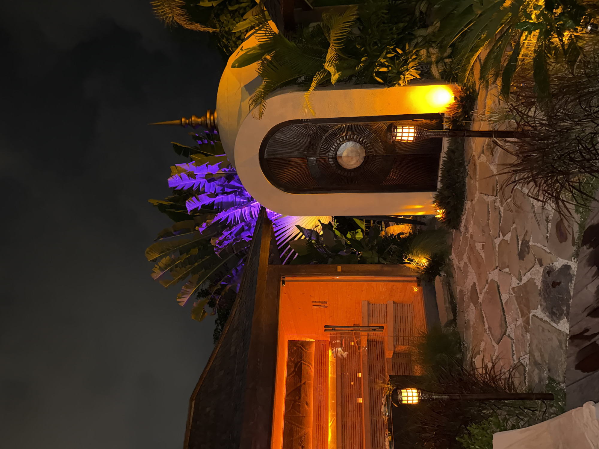The image depicts an outdoor setting at night, likely in Mengwi, Indonesia, given the presence of Balinese architectural elements. A circular structure, possibly a gate or entrance, is centrally located, illuminated with warm light emanating from lamps on either side. Purple lighting accentuates foliage on the left side of the structure, contrasting with the warm light. Another structure with a door is visible below and to the right, also lit with warm light. The ground appears to be paved with stones. The night sky is dark and visible in the background. There are no people visible in the frame.