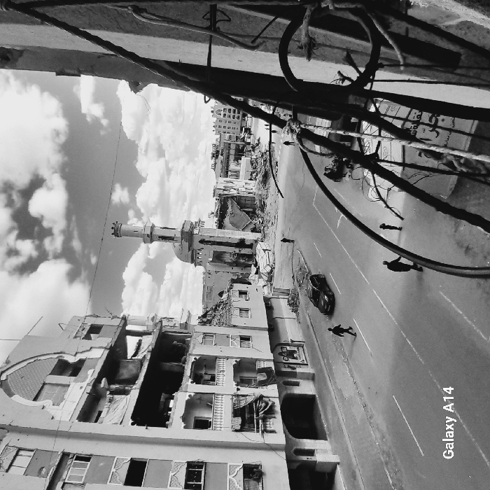 This black and white image, captured from an elevated position in the city of Khan Yunis, Palestinian Territory, depicts a street scene exhibiting widespread structural damage under a sky with scattered clouds. On the left, a multi-story building is severely damaged, with large sections of its facade missing, revealing interior rooms and exposed structural components. A paved street runs along the base of this building. In the midground-center, a mosque-like structure with a domed roof and a tall, cylindrical minaret stands amidst significant rubble and debris from surrounding collapsed or damaged buildings. Further down the street, additional damaged structures are visible in the background. The immediate foreground on the right is partially obstructed by a dense network of twisted rebar and remnants of a building structure, through which a sign with Arabic text is partially visible.
On the street, a dark sedan is positioned diagonally. Several individuals are present: one person walks on the sidewalk on the left, another walks in the middle of the street, two smaller figures are located further right on the street moving away from the viewer, and a fifth figure is visible in the distance near the mosque. The overall environment indicates extensive urban destruction with sparse human presence.