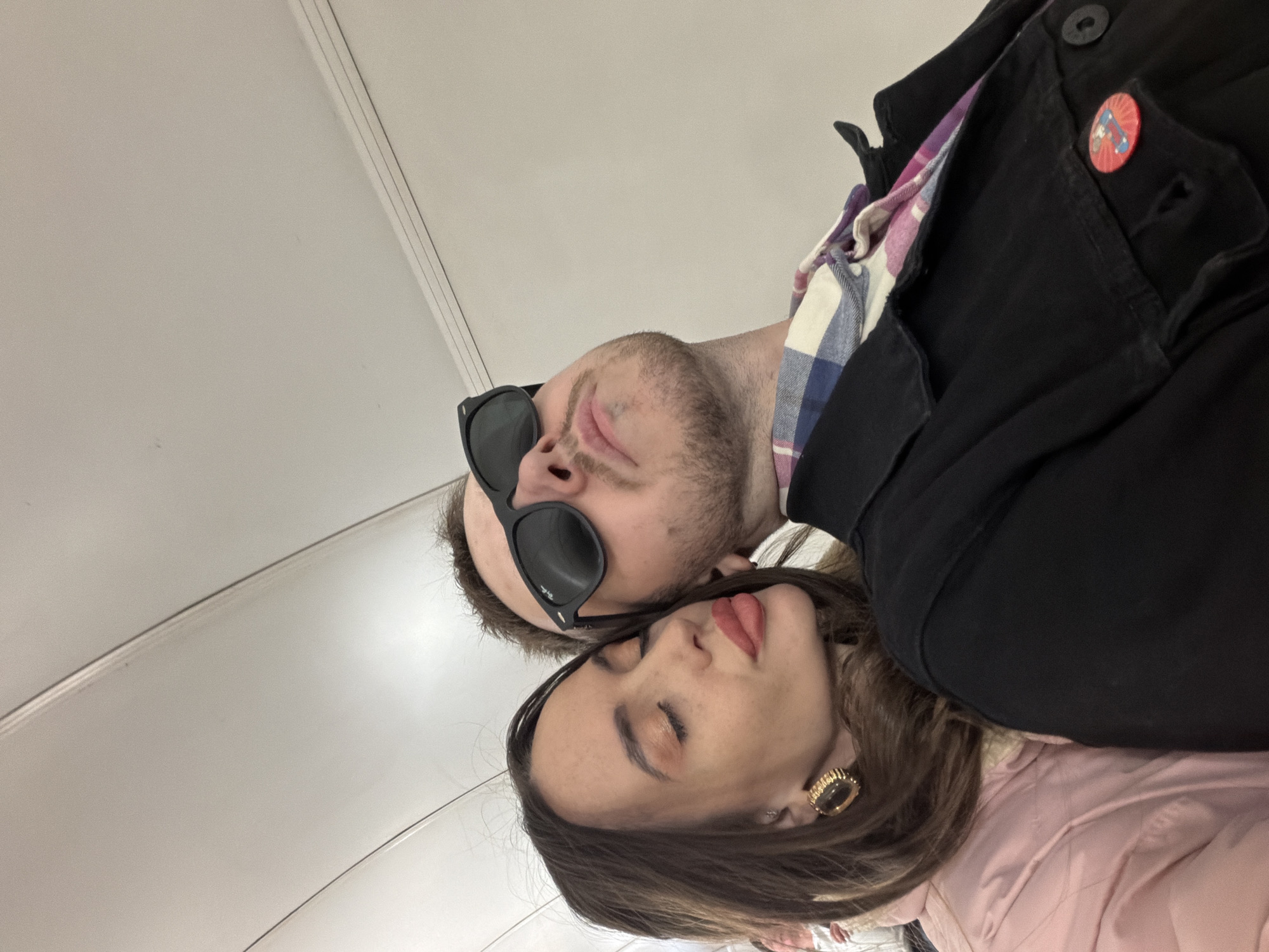 A photograph shows two individuals, a man and a woman, in a close-up shot taken from above. The man is wearing sunglasses, and a black jacket with a pin is visible. A patterned shirt is underneath the jacket. The woman is wearing a pink jacket and a prominent gold earring. The background is a plain, light-colored ceiling. The image does not contain enough information to determine the location within Saint Petersburg, Russia.
