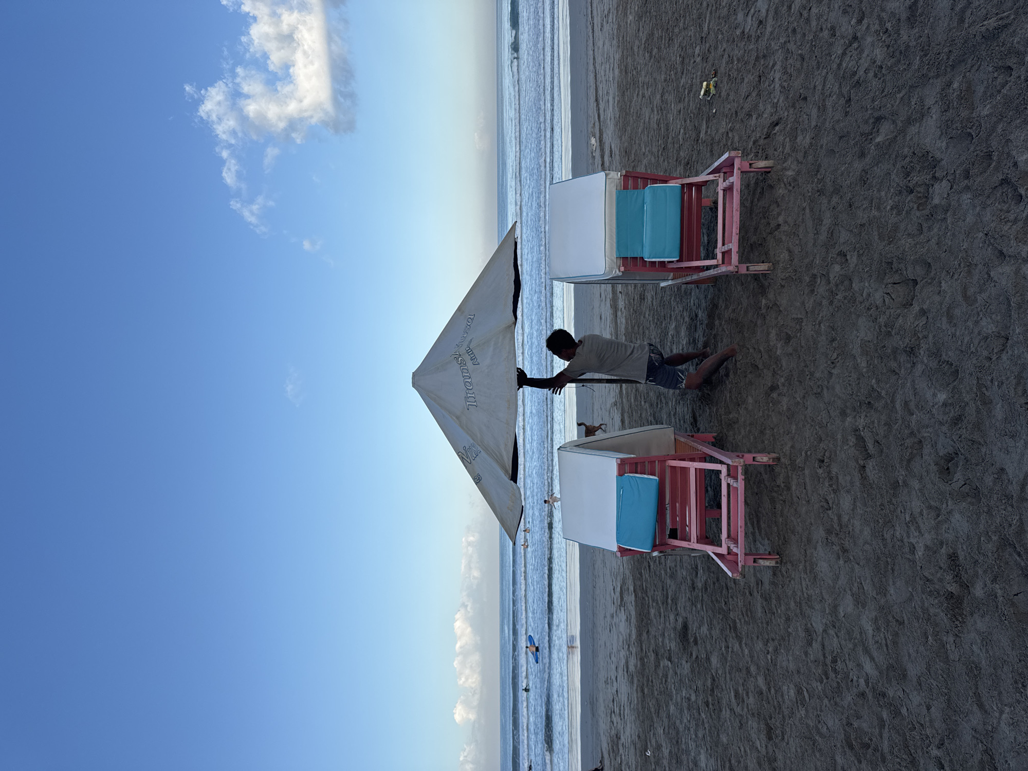 A man erects a beach umbrella in Kuta, Indonesia. The umbrella is white and has text on its side, and two pink beach chairs with blue cushions are near. The beach is sandy, and the ocean is visible in the background. The sky is light blue with a few clouds.