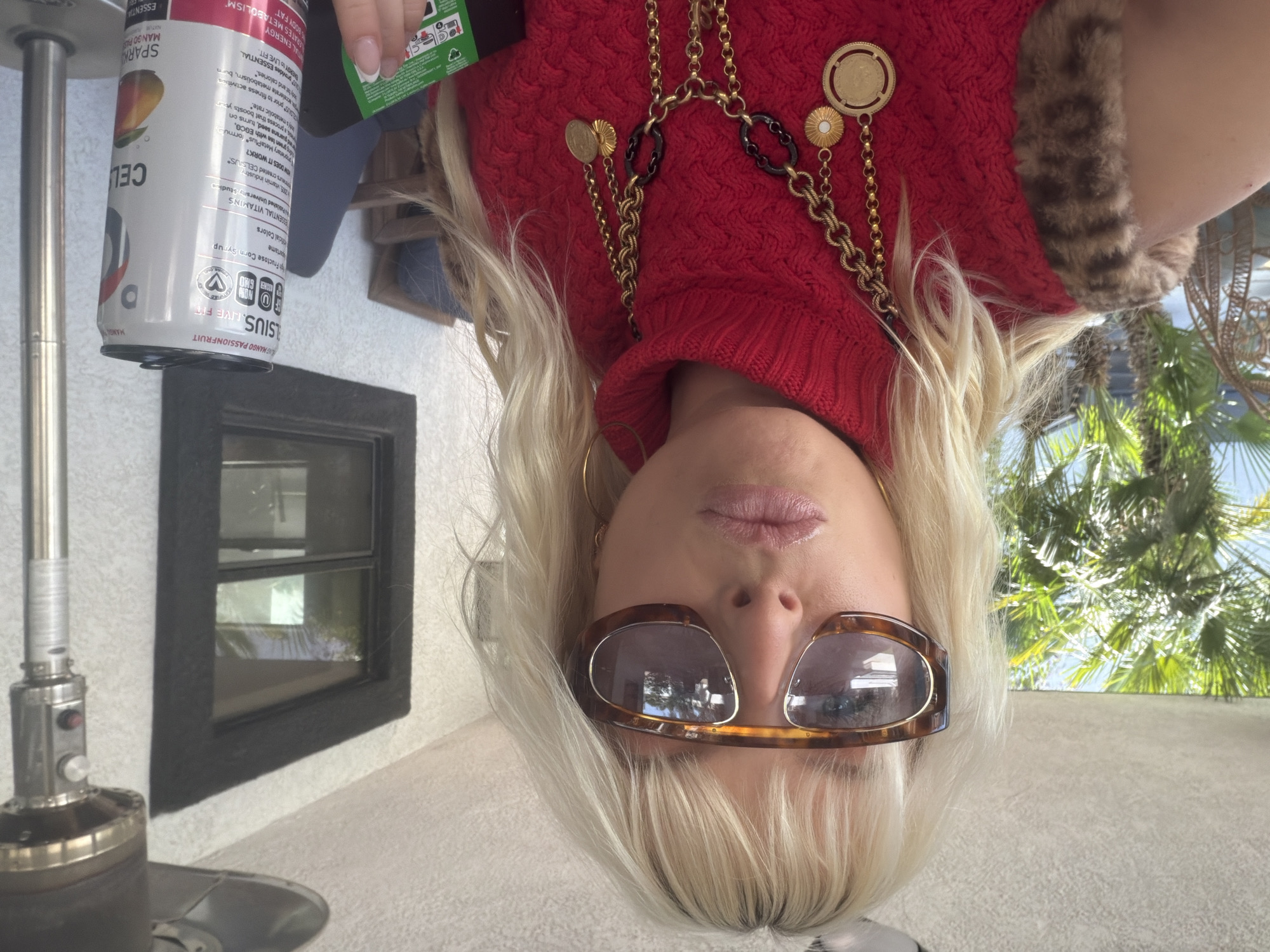 The image, taken upside down, appears to be in Palm Desert, United States. It features a woman with blond hair, wearing sunglasses, a red turtleneck sweater with a fur collar, and gold chains with coin pendants. She is holding a can of Celsius mango passionfruit sparkling drink. There is a patio heater to the left, a black-framed window behind it, and a view of palm trees in the background.