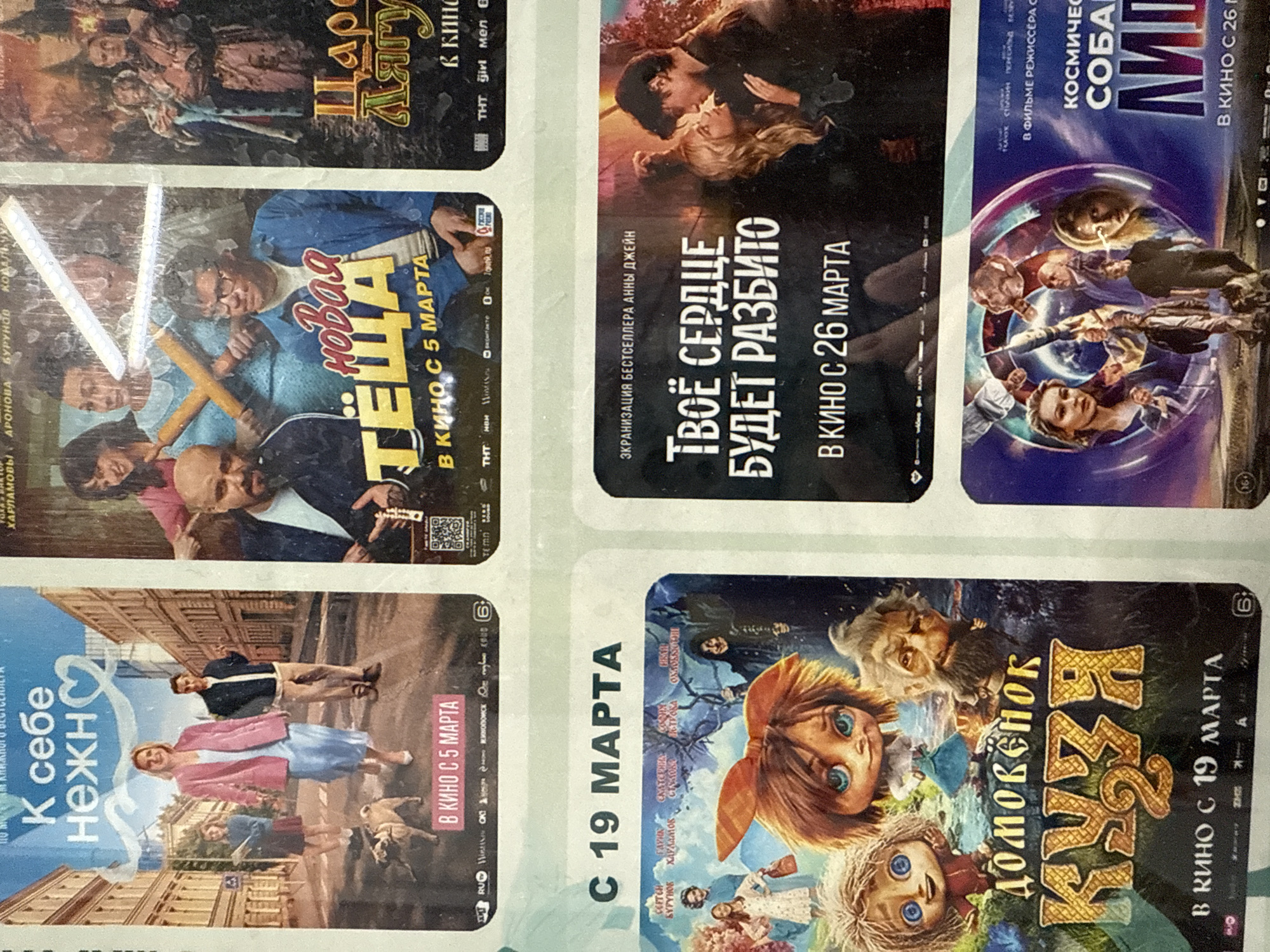 The image shows a close-up of what appears to be a promotional flyer or a display board featuring movie posters. There are six different film posters visible in the image, each in a separate rectangular space, with some overlap.
The movie posters are in Russian and have various titles and release dates, mostly around March. Specifically, release dates like "March 5", "March 19", and "March 26" are visible.
Based on the information provided in the surrounding text, the image was taken in Votkinsk, Russia.