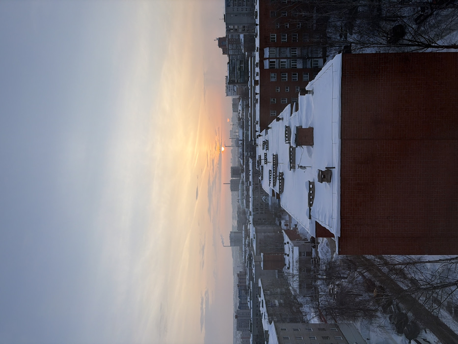 The image displays a panoramic view of a snow-covered urban landscape in Izhevsk, Russia, captured at either sunrise or sunset. The sky, occupying the upper portion, transitions from a pale blue overhead to warm orange and yellow hues near the horizon, where the sun is visible as a bright disc, partially obscured by distant structures. Thin, scattered clouds are present.
In the foreground, the flat, snow-covered roof and the upper section of a red-brick multi-story residential building are prominent on the right. This roof features multiple red-brick chimneys, several smaller square, snow-covered structures, and a satellite dish. To the immediate left of this building, a snow-covered street or access road is visible, lined with bare trees and several parked vehicles.
The midground and background present a dense urban sprawl consisting predominantly of numerous multi-story residential apartment buildings with light-colored facades, all topped with a substantial layer of snow on their roofs. Interspersed among these buildings are bare trees. In the distant background, industrial elements are visible, including multiple smokestacks, some emitting plumes of white vapor. No individuals, explicit actions, or interactions are discernible within the scene. The pervasive snow cover and visible atmospheric conditions indicate a cold, clear winter day.