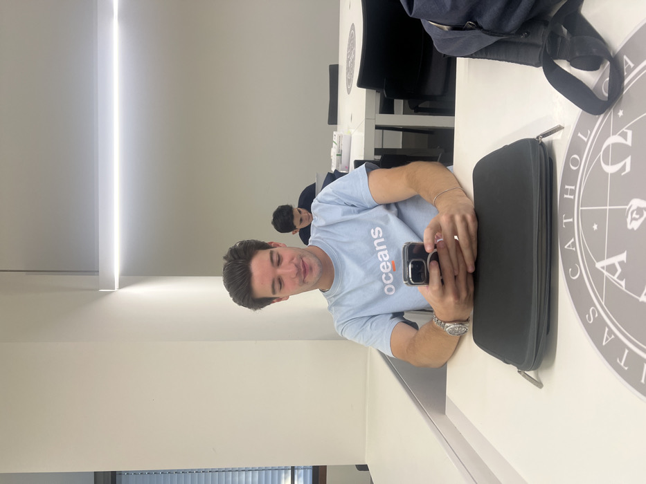 This image depicts an indoor scene within the Universidade Católica Portuguesa - Porto Campus, located in Foz do Douro, Aldoar, Foz do Douro e Nevogilde, Portugal.
In the foreground, a young adult male with dark brown hair and light skin is seated at a white table. He is wearing a light blue t-shirt with the word "oceans" and an orange horizontal line printed on the chest. On his left wrist is a silver wristwatch, and on his right wrist is a thin silver bracelet. He holds a dark-colored smartphone, equipped with a back grip accessory, in both hands, looking down at its screen with a slight smile. On the table in front of him, a black zippered case is visible, positioned next to a circular grey mat displaying the "UNIVERSITAS CATHOLICA" emblem. A dark-colored bag with a visible strap is partially present on the far right edge of the table.
In the mid-ground, another young adult male with dark hair is seated at a white table, positioned behind and to the right of the foreground subject. He wears a dark top and is looking down at a laptop. On his table, a white box and a stack of white rectangular items are present. Black office chairs with armrests are situated around this table.
The room features plain, light-colored walls and is illuminated by a prominent, linear LED light fixture mounted horizontally on the ceiling. A window with horizontal blinds is partially visible on the left side of the frame. The overall environment appears to be a modern, well-lit study or common area.
