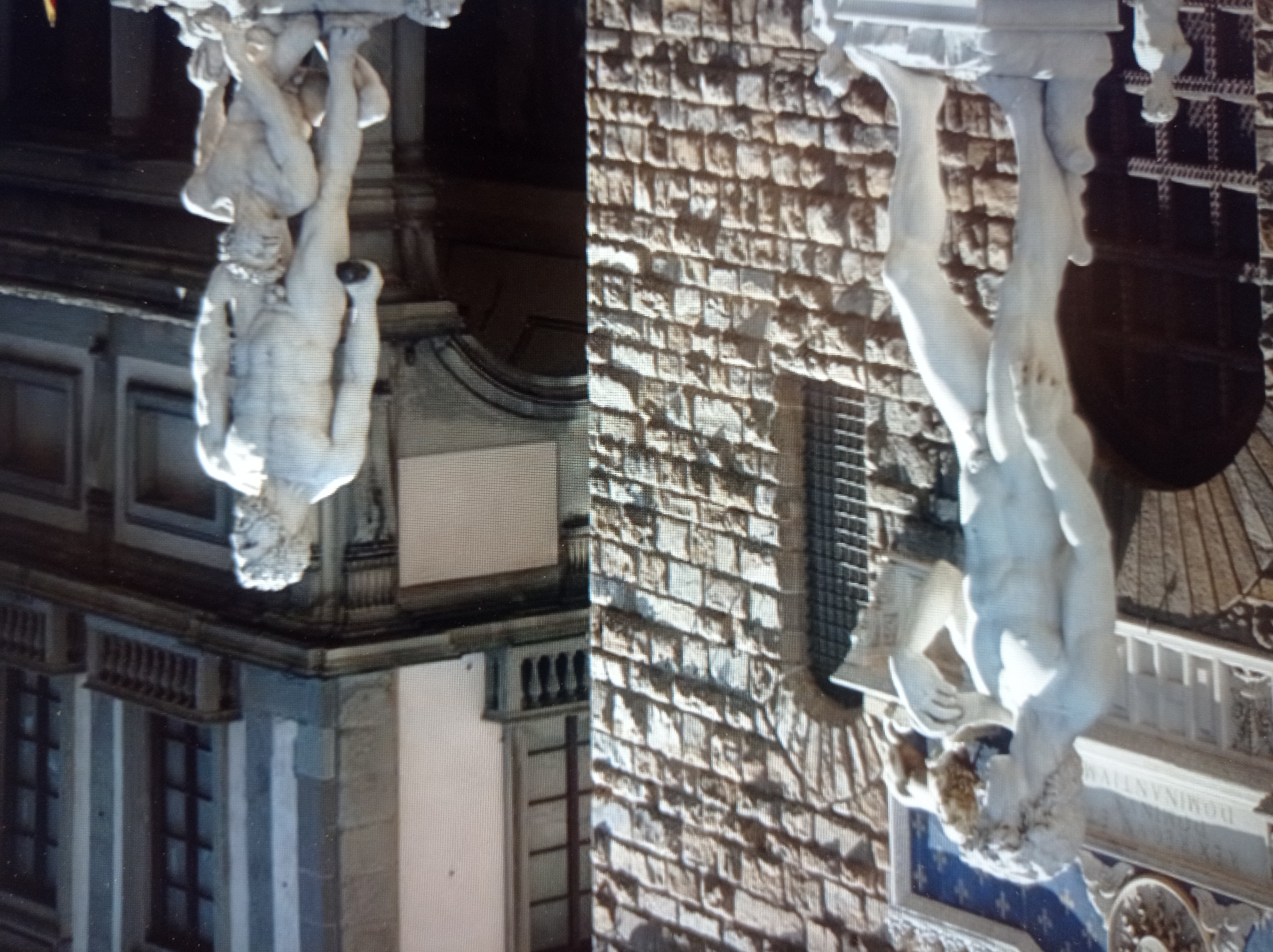 The image depicts a plaza with classical sculptures in front of a stone building. On the left, a large white marble statue of a male figure with curly hair stands with his right arm extended, holding a stone. He is positioned in front of a textured stone wall with an arched entryway and a barred window. To the right, a larger white marble sculpture features a muscular male figure standing over a smaller crouching male figure. The standing figure is holding a round object. The building in the background has multiple stories with windows and balconies. The overall scene suggests an outdoor public space adorned with historical art. There is no indication that this location is in Mukachevo, Ukraine.