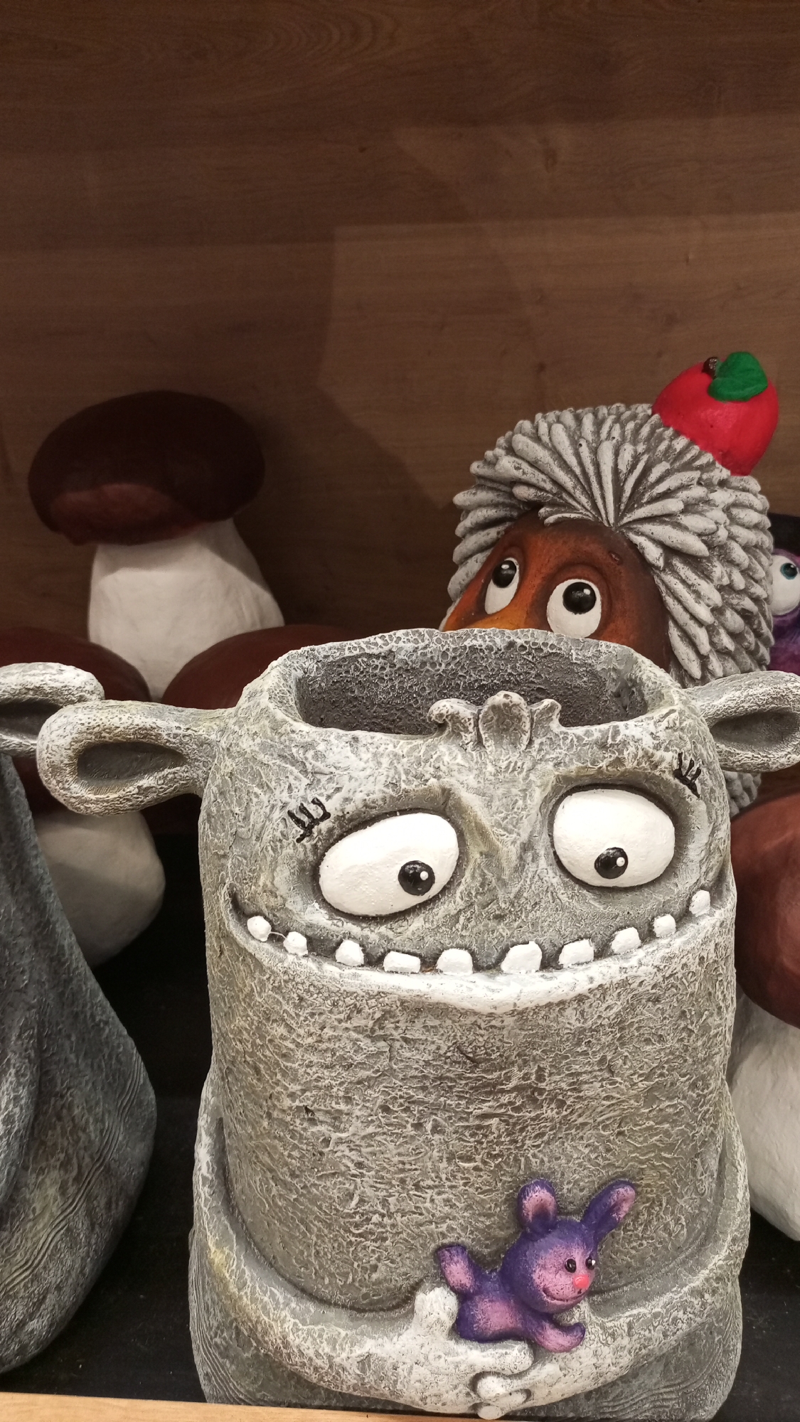 The image shows a close-up of a gray, textured ceramic pot with a whimsical monster face. The pot has large, cartoonish eyes, a toothy grin, and ear-like protrusions on the sides. A small, purple rabbit figurine is attached to the lower portion of the pot. In the background, there are several decorative ceramic mushrooms and a gray-haired gnome-like figure with an apple on its head. The background appears to be a wooden shelf, suggesting the setting is a retail environment. There is no information or indicators suggesting the image was taken in the city of Yudino, Russia.
