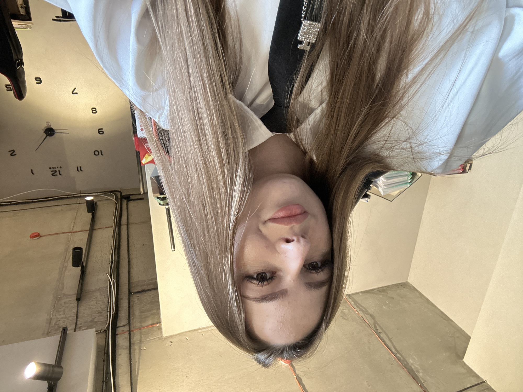 An indoor selfie shows a young woman with long, blonde hair, wearing a white collared shirt and a black tie with a silver charm. She is smiling and looking at the camera. In the background, the ceiling is exposed concrete with pipes and lighting fixtures. A large wall clock with Roman numerals is visible on the right side of the frame. The environment appears to be an office or workspace.