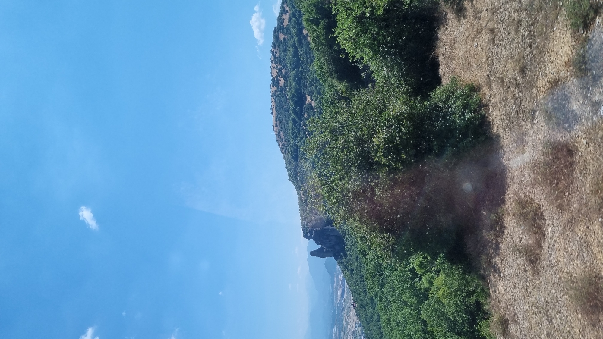 The image shows a view of the Meteora rock formations and the surrounding landscape near Kalambaka, Greece. In the foreground, there is dry, rough terrain with sparse vegetation. A dense forest of green trees occupies the middle ground, with a prominent rock formation, characteristic of Meteora, rising from the trees on the left side of the frame. The rock formation consists of several tall, weathered stone pillars. Beyond the forest and rocks, a valley with scattered buildings and further mountain ranges are visible in the distance. The sky above is clear blue with a few wispy white clouds. The overall impression is a natural landscape with a distinct geological feature.