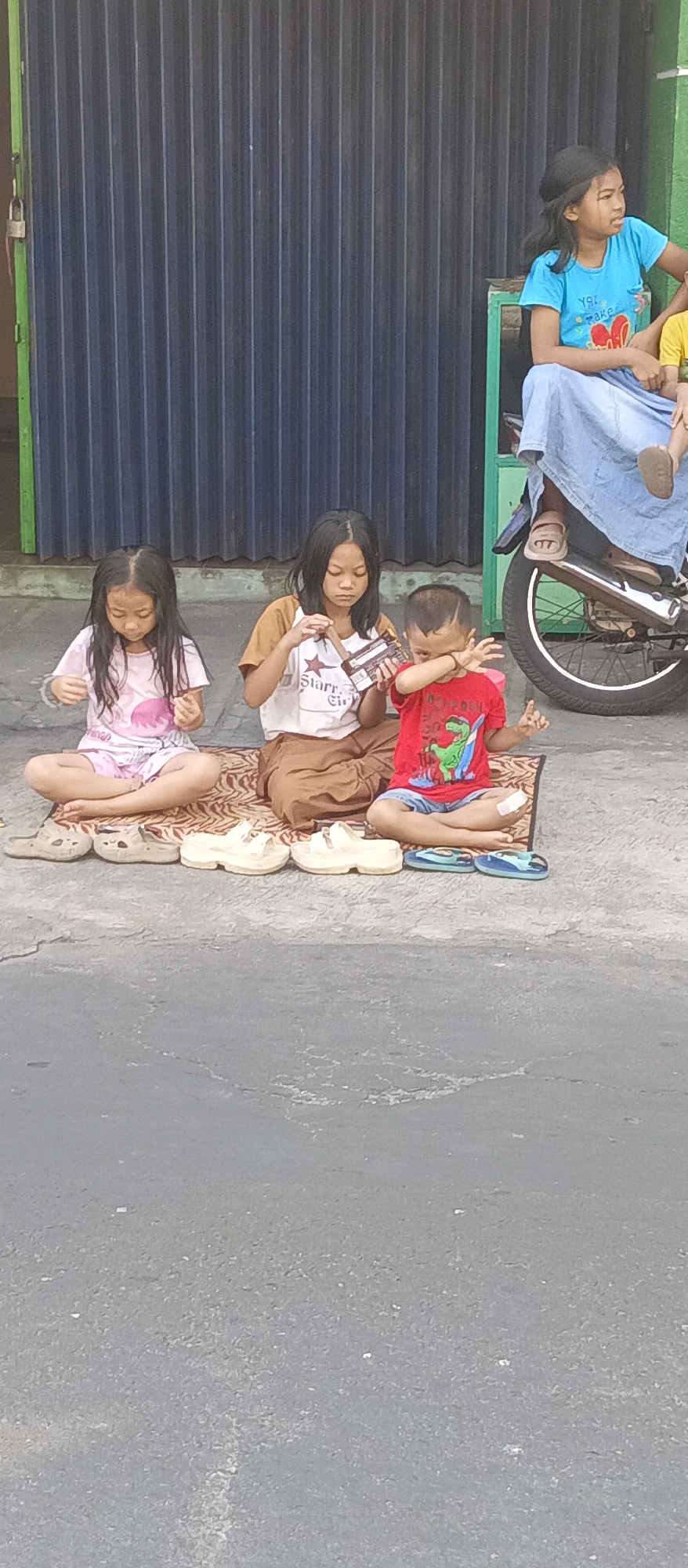 In Malang, Indonesia, four children are seated on a mat on a paved surface in front of a dark, corrugated metal shutter. The shutter appears to be part of a storefront or building facade. To the right, a motorcycle is parked, with a person wearing a blue shirt and a light blue skirt or wrap seated on it, holding a young child.
Three children are seated directly on the mat. From left to right:
* The first child, a girl with long dark hair, wears a light pink t-shirt with a white and pink graphic. She is looking down at something in her hands.
* The second child, a girl with long dark hair, wears a brown and white t-shirt with the words "Star Sir" visible. She is holding a chocolate bar wrapper and appears to be examining it.
* The third child, a young boy with short dark hair, wears a red t-shirt with a green dinosaur graphic and blue shorts. He has his right hand covering his eyes and his left hand is raised. A bandage is visible on his right knee.
Arranged in a row in front of the children on the mat are several pairs of sandals.
The environment suggests an outdoor public space, likely a sidewalk or street in front of a commercial establishment. The time of day appears to be daytime, given the lighting and shadows.