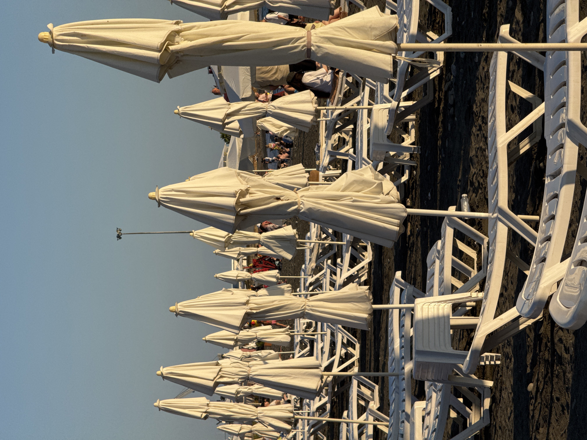 The image depicts a beach in Sochi, Russia, likely during the late afternoon or early evening, as indicated by the warm, low-angled sunlight casting long shadows. The foreground is dominated by rows of tightly packed white plastic lounge chairs and closed white beach umbrellas. The chairs are mostly empty, arranged on dark, pebbly sand. In the mid-ground and background, the beach appears more populated with people sitting and standing, some near the water's edge which is not visible in this frame. The umbrellas are all closed, their fabric taut and secured by ties. Some of the umbrellas have yellow caps on top. A lamppost with multiple lights is visible in the distance, near the horizon. The sky is clear and a light blue, transitioning to a warmer tone near the horizon. The overall impression is one of a beach preparing to close for the day, with lounge chairs and umbrellas neatly stored.