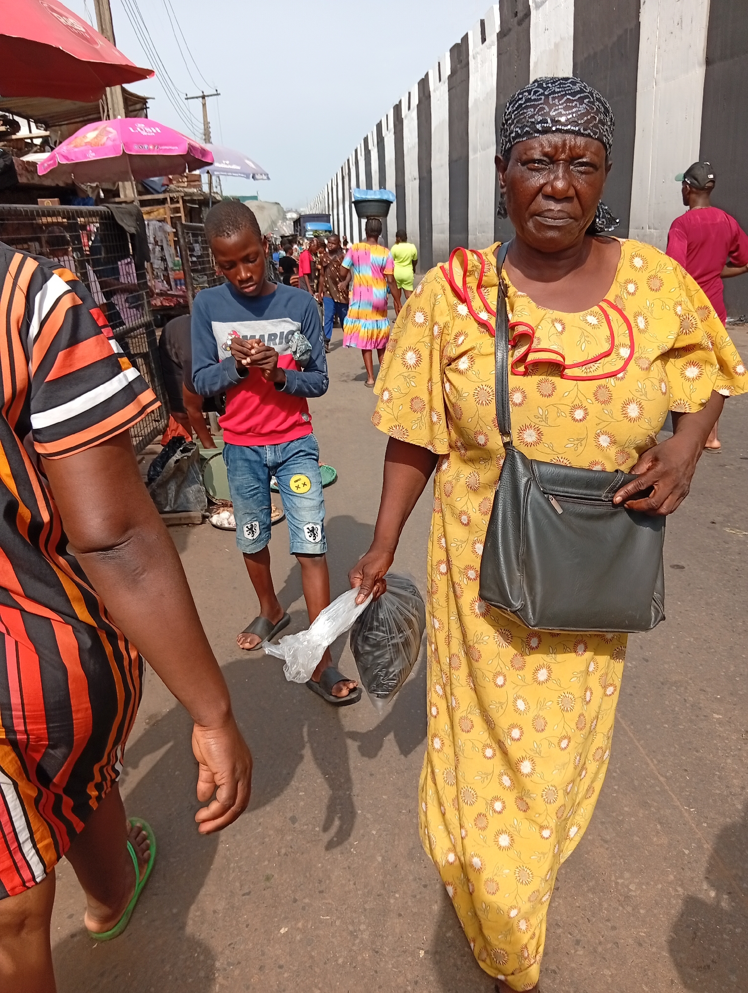 The image depicts a street scene in Nkpor, Nigeria. An older woman in a long yellow dress with a floral pattern and red trim is walking towards the viewer. She has a black headscarf and is carrying a black purse on her shoulder and a plastic bag in her hand. A young boy is seen standing in the background, looking at something in his hands. Other people are walking around in the background. A concrete structure with vertical black and white stripes is visible in the background. Market stalls with colorful umbrellas line the street. The ground is paved.