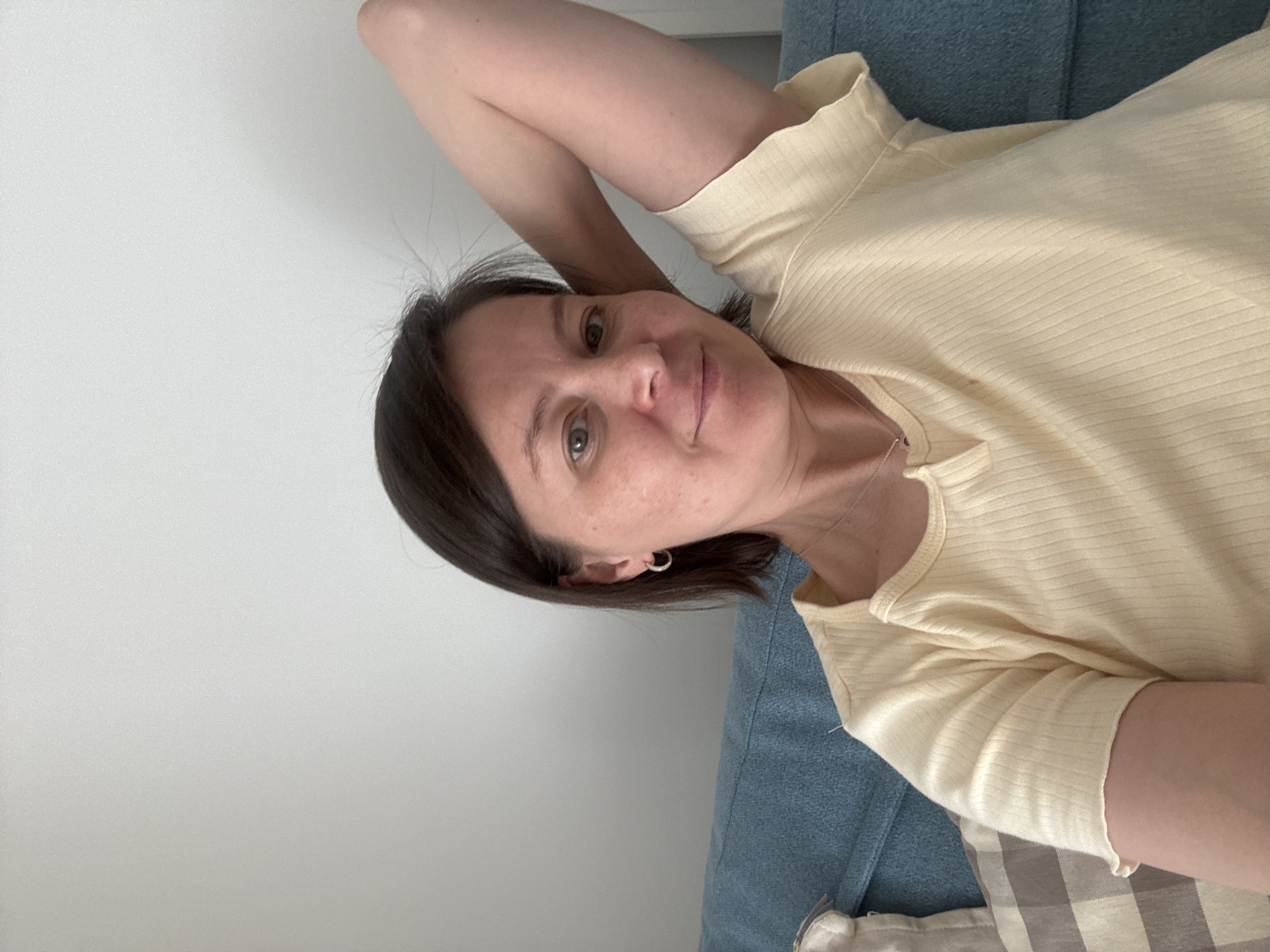 The image is a rotated, close-up portrait of an adult woman in a casual indoor setting. She is lying down, resting her head on her left arm, and looking directly at the camera with a gentle, relaxed smile. She has short, dark brown hair, fair skin with visible freckles, and light-colored eyes.
The woman is wearing a light yellow, ribbed short-sleeved top with a subtle V-neckline. A thin silver necklace is visible around her neck, and she has a small silver hoop earring in her left ear.
The background consists of a plain, light-colored (likely white or off-white) wall to the left and above her. A textured blue fabric, possibly part of a sofa or bed, is visible to her right and below. A small section of a checkered pattern fabric, likely a pillow or blanket, can be seen in the lower right corner. The lighting is soft and even, suggesting an indoor daytime setting. The overall mood conveyed is calm and personal, likely a selfie taken in a home environment in Izhevsk, Russia. No visible text is present in the image.