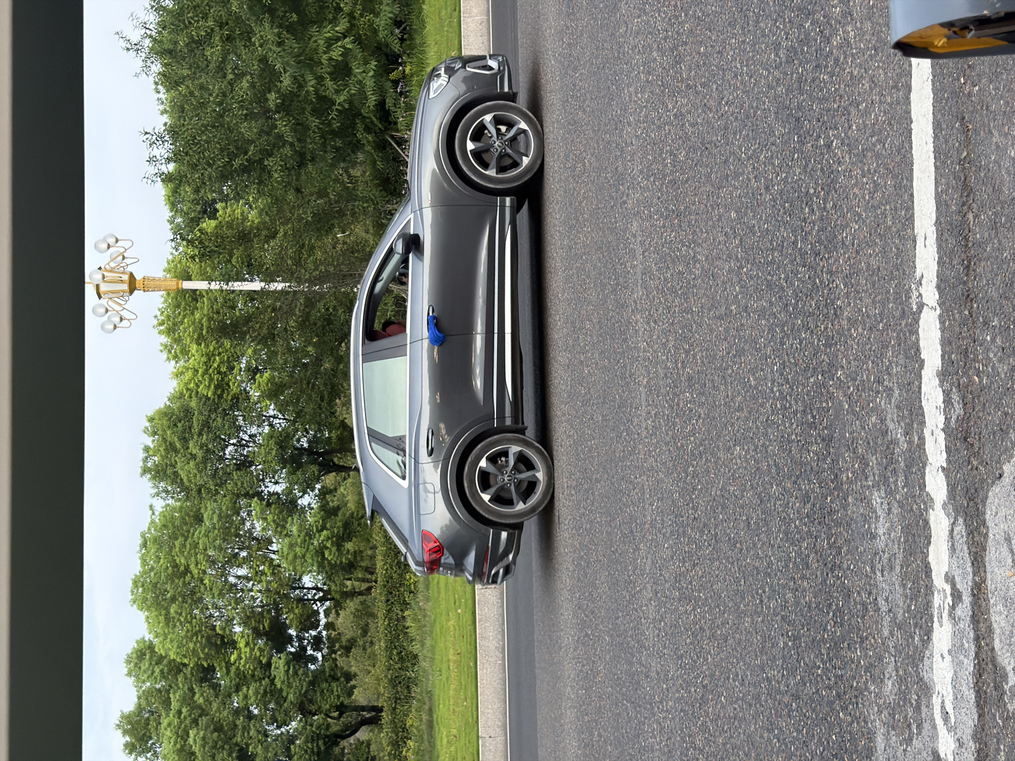 A dark grey Audi SUV is driving on a paved road in Jiangjin, China. The vehicle is captured in a side profile shot, showing its sleek design and alloy wheels. A blue cloth is attached to the driver's side door handle, possibly as a marker or decoration. The background consists of a row of lush green trees and a cloudy sky. A decorative lamppost with multiple globes stands tall among the trees. The road surface is asphalt with a white line painted on it, indicating a lane divider. The lower portion of the image shows a grey and white striped marker on the edge of the road.