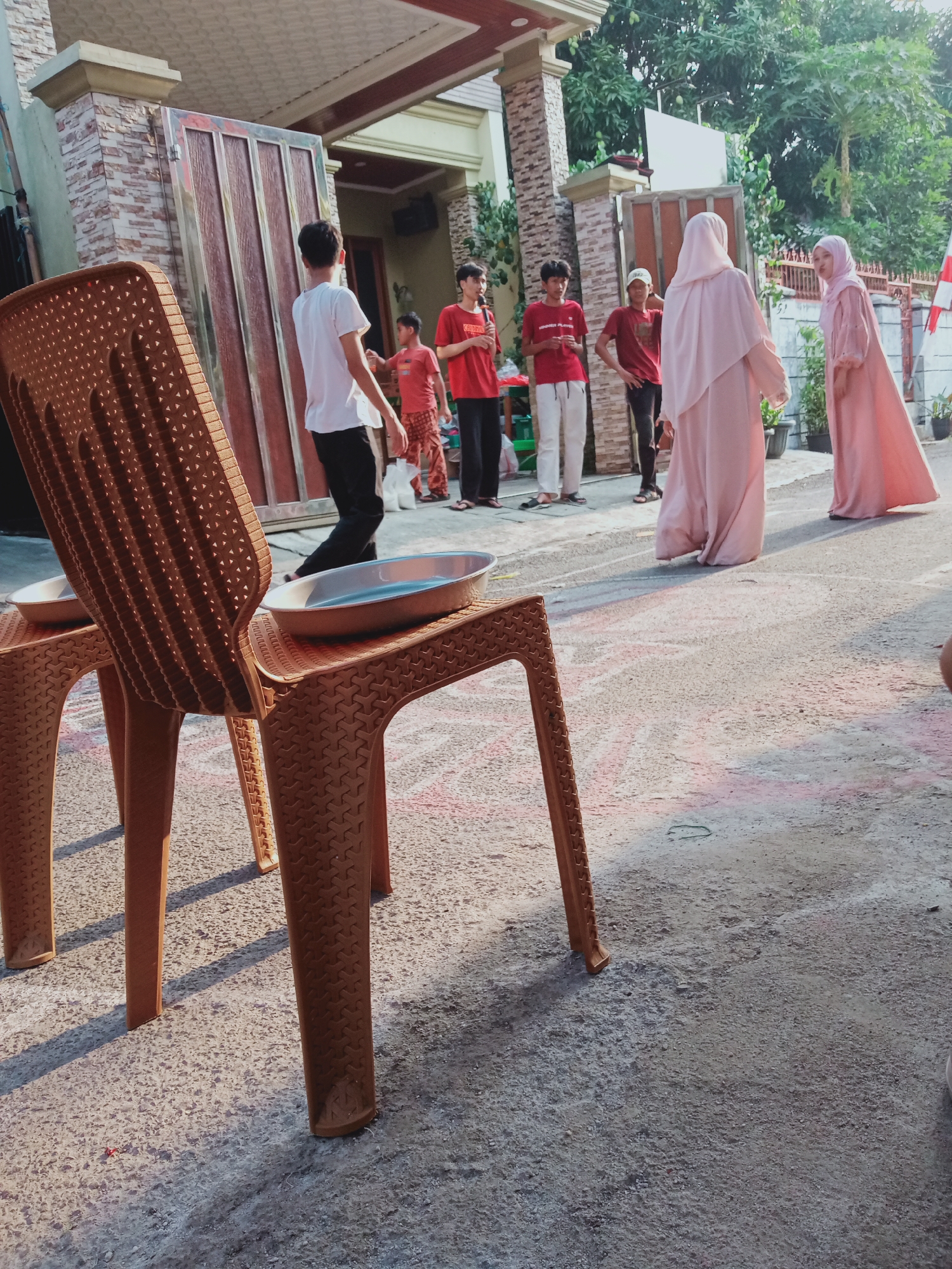 In East Jakarta, Indonesia, on a paved street with chalk drawings, a group of people are gathered in front of a house with a stone facade and a large wooden gate. In the foreground, two brown plastic chairs are positioned, with metal basins resting on them. A young boy in a red shirt and patterned pants stands near a table. Several young men are also present, some wearing red shirts and white pants, others in a white t-shirt and black pants. Two women, dressed in pink hijabs and long dresses, are facing each other and interacting. The scene appears to be a casual outdoor gathering.