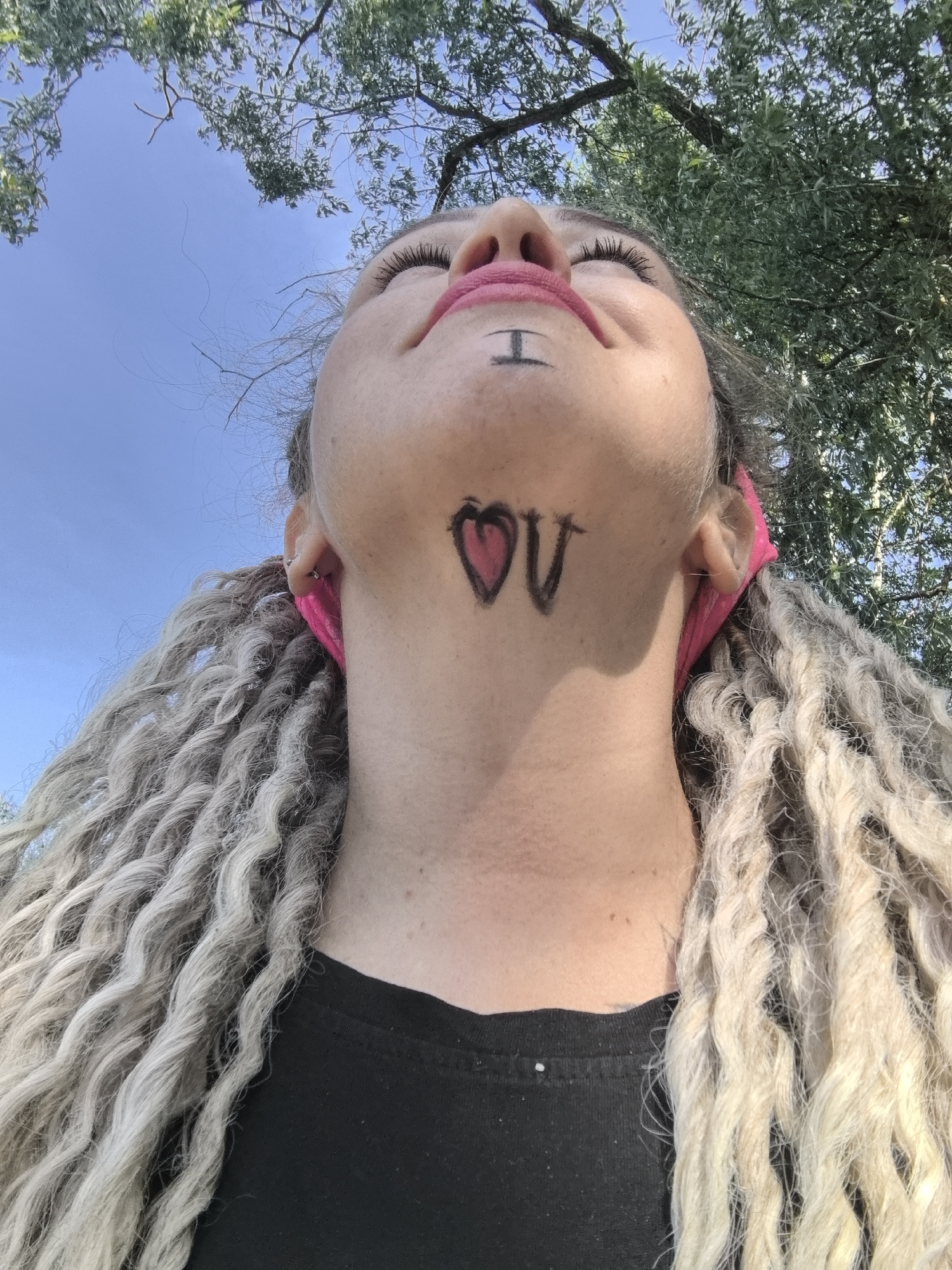 A woman with silver, rope-like braids is shown from a low-angle perspective, with a background of trees and a blue sky. The woman has an "I" drawn under her lower lip and a heart and "U" drawn on her neck. The woman is wearing a black shirt. The location is not specified.