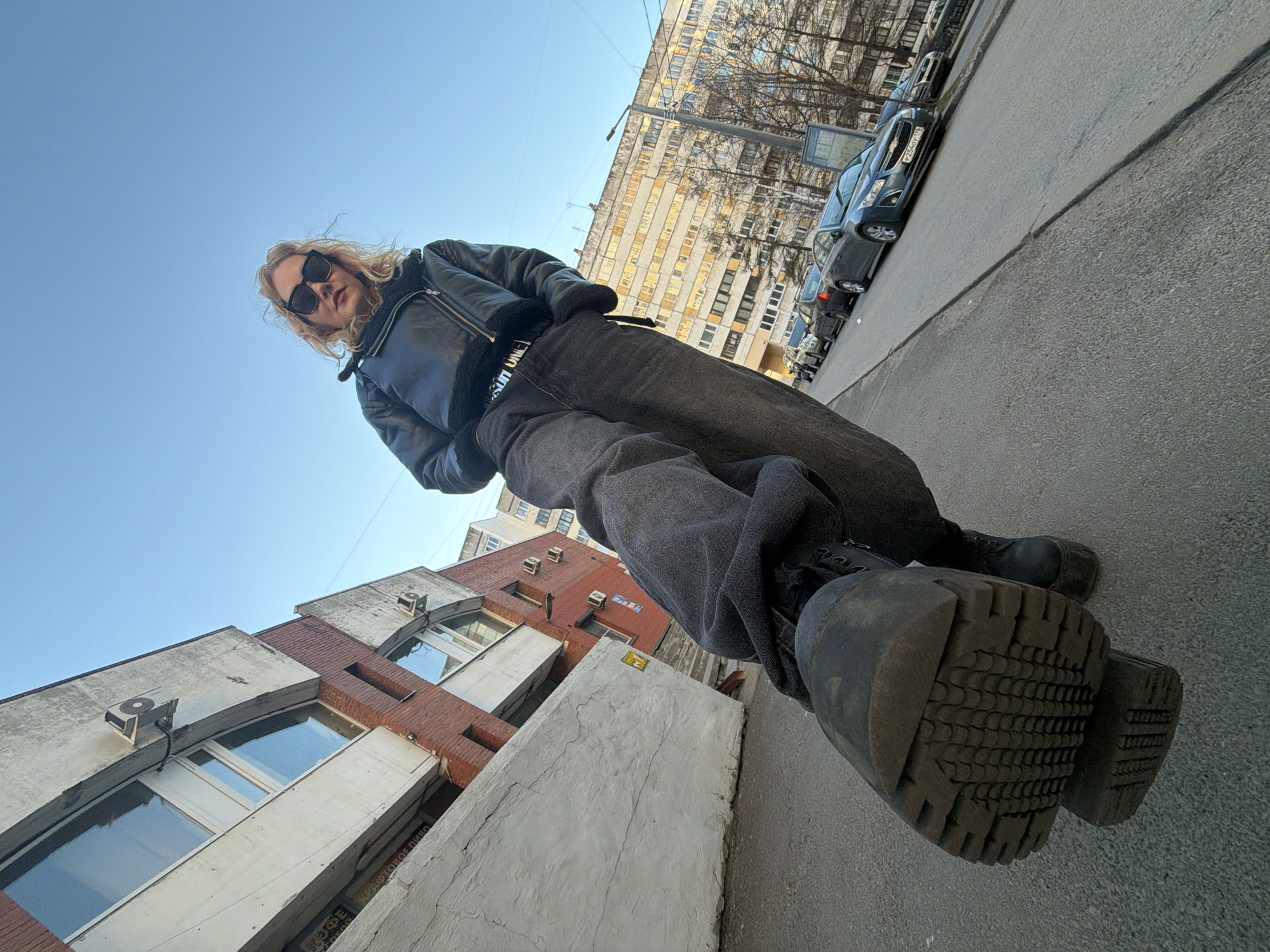 A low-angle shot shows a person in Saint Petersburg, Russia, standing on a sidewalk next to a street. The person wears black sunglasses, a black leather jacket, dark-colored jeans, and black boots with thick, textured soles. In the background is a multi-story building with a mix of white and red brick sections, and windows. Air conditioning units are visible on the building's exterior. Beyond the building, there is a street with parked cars. The sky above is clear and blue. The photograph appears to have been taken with a wide-angle lens, giving a distorted perspective.