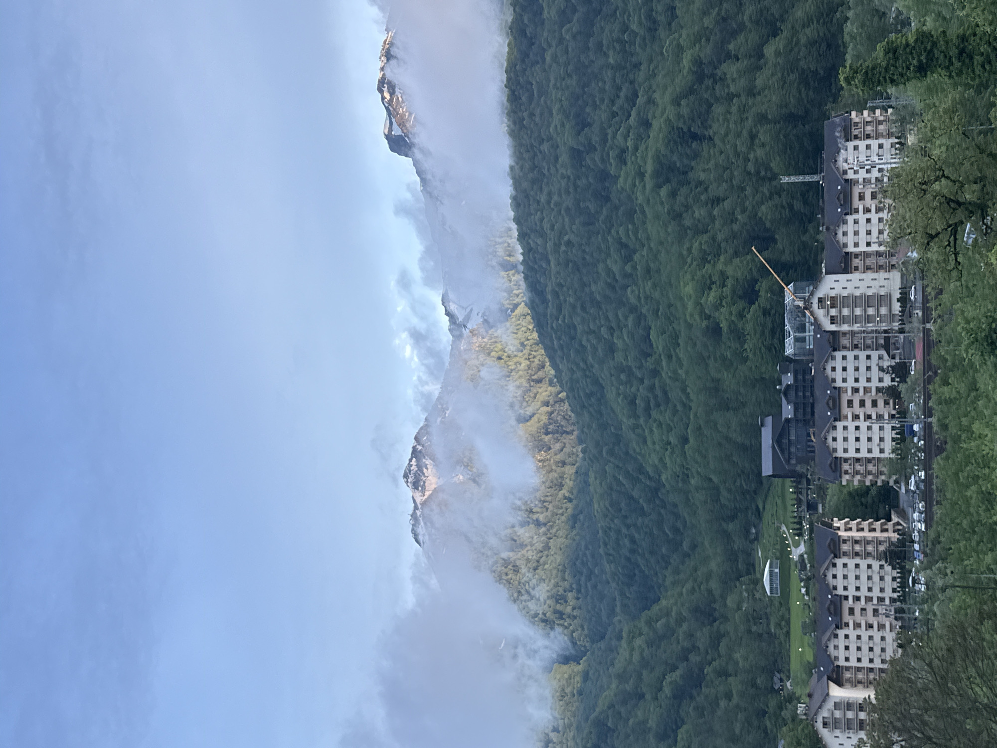 The image shows a view in Krasnaya Polyana, Russia. Mountains are shrouded in clouds, a dense green forest covers the slopes and at the base of the mountain are clustered buildings, seemingly hotels or apartments. A construction crane is visible among the buildings.