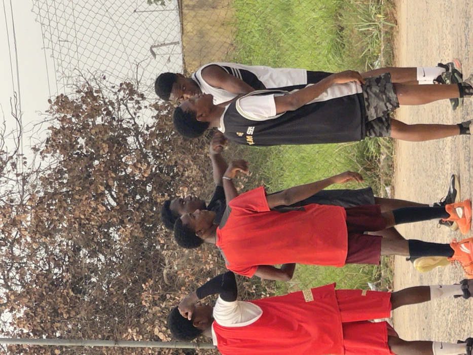 The image captures a group of six young males outdoors, appearing to be engaged in a sporting activity or warm-up. They are all standing and looking upwards, some with their arms slightly raised or positioned near their heads, suggesting they might be stretching, watching something above them, or mimicking a specific movement.
The individuals are dressed in athletic wear. In the foreground, one person wears a red short-sleeved shirt with a white design on the left shoulder and red shorts, with black sleeves or bands on their forearms and dark shoes with white socks. Next to him, another person is in a red short-sleeved top and maroon shorts, wearing black socks and bright orange athletic shoes. Behind them, another individual wears a dark short-sleeved top and dark shorts. Further back, a person is seen in a black sleeveless jersey with white trim and a white undershirt, paired with camouflage-patterned shorts, white socks, and dark athletic shoes. Part of the text "THIS W" is visible on his black jersey. Behind him, another person wears a white sleeveless jersey with black side stripes and dark shorts, dark socks, and dark athletic shoes. A sixth person is partially visible behind the group.
The setting is an outdoor area during the daytime. The ground beneath them appears to be a mix of dirt or a light-colored paved path, transitioning into green grass or low vegetation in the middle ground. A chain-link fence stretches across the background, and behind it, there are trees with numerous small, brown, dry-looking leaves, contrasting with the green grass. The sky above is bright, suggesting an overcast day or simply bright daylight. The overall impression is that of a practice session or casual gathering in an open, possibly semi-urban or rural environment.
