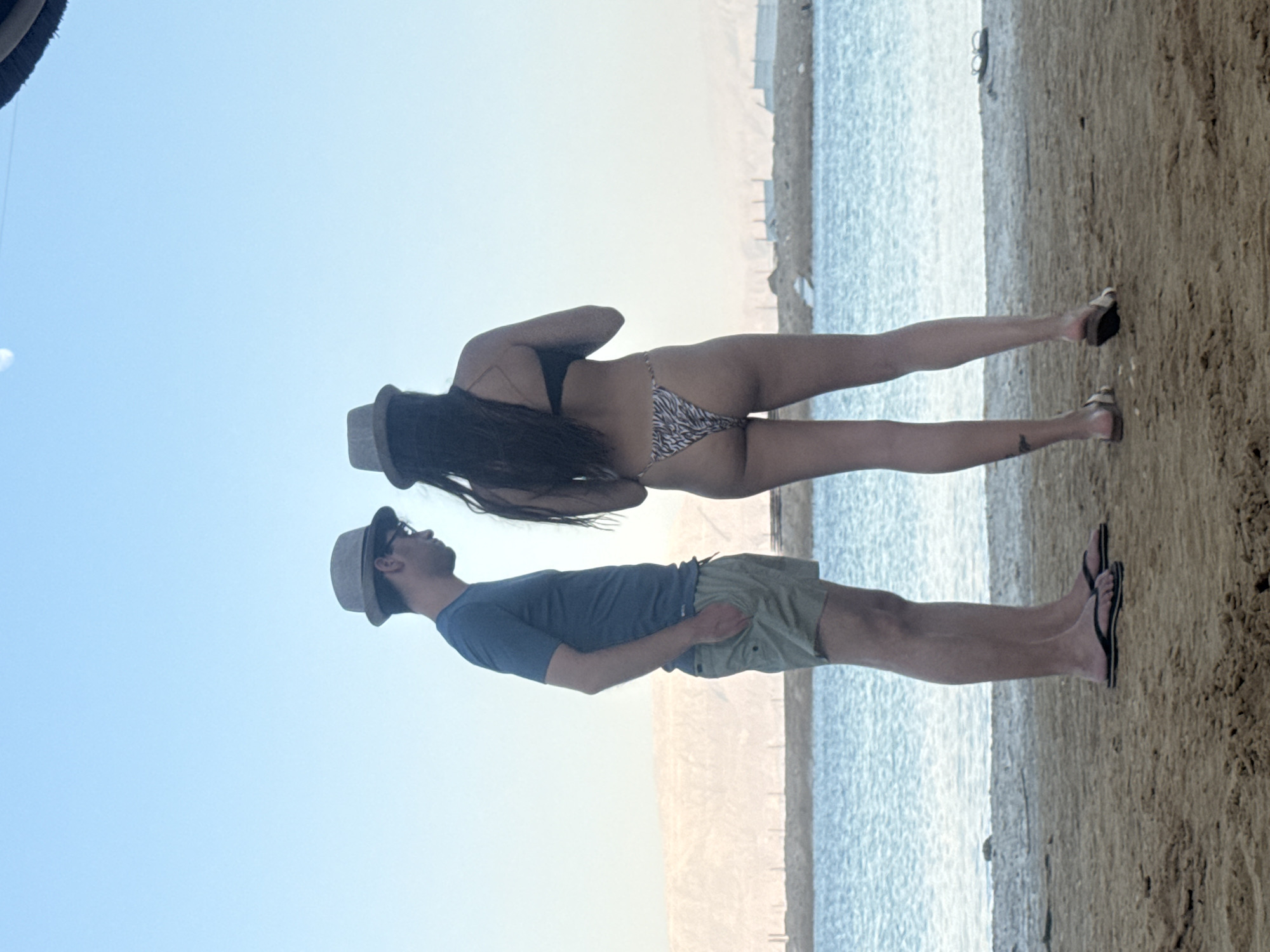 Two individuals, a man and a woman, are posing on a beach. The man, wearing a blue t-shirt, light green shorts, and a hat, stands with his back to the camera, looking out at the water. The woman, in a bikini and a hat, is positioned behind him, facing the viewer. She has long, dark hair and is leaning slightly forward.
The scene is set on a sandy beach with the ocean to the right. The water appears calm and the sky is a clear, light blue, suggesting a bright, sunny day. The sand is light brown and textured. In the distance, there's a hint of a shoreline or a man-made structure. The overall impression is one of relaxation and leisure. No text is visible in the image. The orientation of the image is portrait.