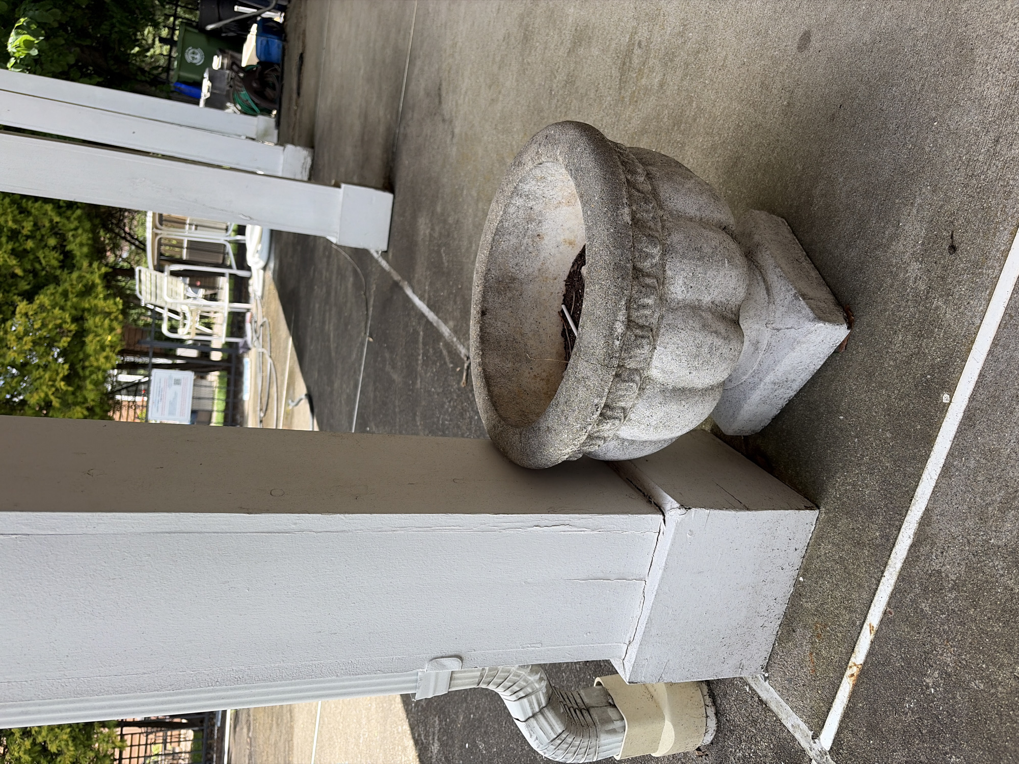 A partially obscured, medium shot depicts an exterior location in Edison, United States. The scene prominently features a weathered, gray stone or concrete urn resting on a painted white wooden beam. The urn sits slightly askew, revealing some internal debris or dried plant matter. The beam is adjacent to a white, rectangular column, likely part of a porch or patio structure. A white downspout runs along the bottom of the beam. The concrete surface appears well-worn and gray, with subtle surface variations. In the blurred background, there are glimpses of greenery, white patio furniture, and a green trash container, suggesting a residential or commercial outdoor space.
