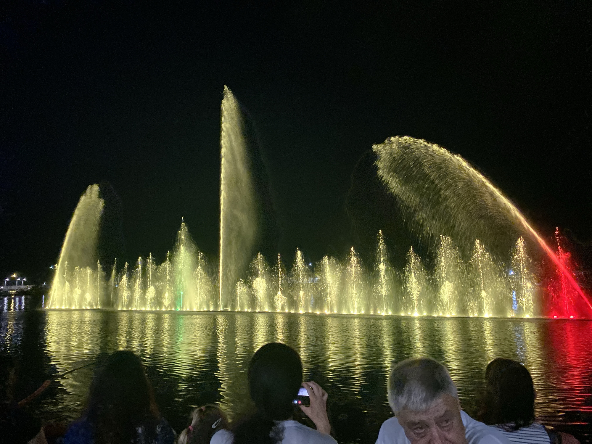 A long exposure photograph captures a nighttime view of a synchronized water and light show on the Pomorie Lake in Pomorie, Bulgaria. The dark sky is illuminated by numerous illuminated water jets, creating a dynamic display of cascading water lit in yellow, green, and red hues. The light reflects vibrantly on the dark, rippling surface of the lake. In the foreground, partially obscuring the view of the water show, are the backs of several people. A woman in the center foreground is holding a small camera to her face, seemingly taking a photograph of the spectacle. To her right, an older man with gray hair is visible, looking forward. The scene depicts a public attraction and leisure activity.