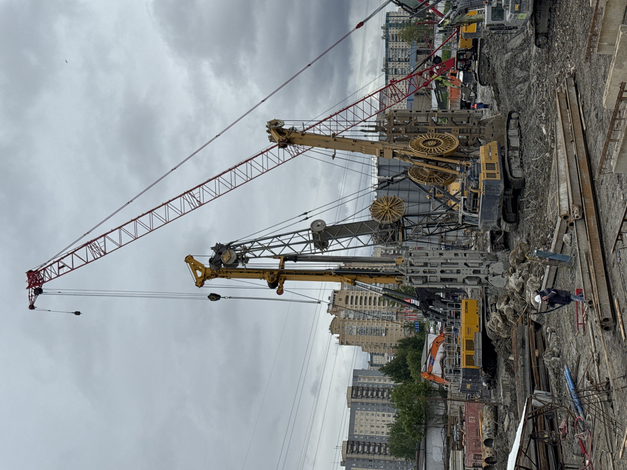 The image depicts a construction site in Saint Petersburg, Russia. Several cranes and drilling machines are visible, with a red crane boom extending high into the cloudy sky. Buildings are visible in the background, suggesting an urban setting. Construction workers are present near the machinery, amidst a muddy environment with exposed metal rebar and pipes.