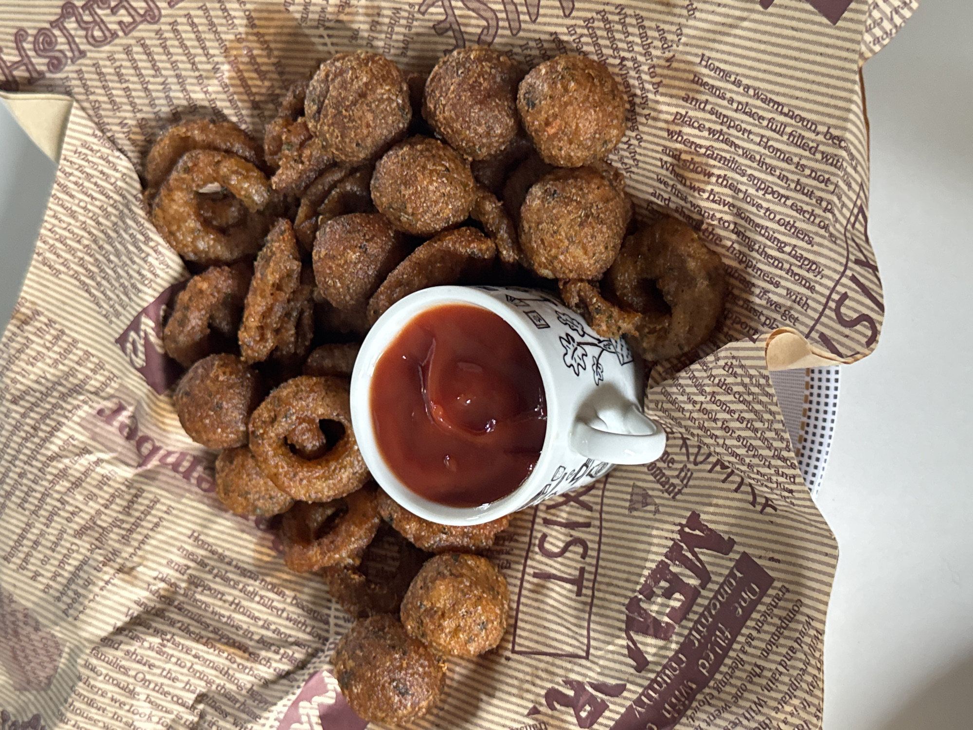The image displays a close-up, overhead view of a snack served in a crinkled paper lining, resembling a basket. The main subjects are two types of fried food: numerous golden-brown, ring-shaped items, possibly onion rings, and several small, round, similarly fried items, which could be meatballs, hushpuppies, or similar savory bites. In the center, a small white ceramic cup with a delicate black pattern holds a serving of bright red ketchup, acting as a dip for the fried snacks.
The paper lining is printed with text in a newspaper or journalistic style. Prominently visible is a paragraph defining "Home" as "a warm noun, because means a place full filled with love and support. Home is not just a place where we live in, but a place where families support each other and show their love to others. When we have something happy, we first want to bring them home and share the happiness with families. On the contrary, if we get into trouble, home is the first place where we look for comfort." Other fragmented words like "Remember," "patience," "prosperity," "restrained," "mood," "MEMOR," and "KYST" are also discernible.
The scene suggests a casual dining or take-out experience, likely a snack or appetizer. No people are visible, nor are there any specific time-of-day or weather indicators. While the location context provided is Davao City, Philippines, the image itself does not contain any explicit visual cues or landmarks to confirm this specific geographical setting.
