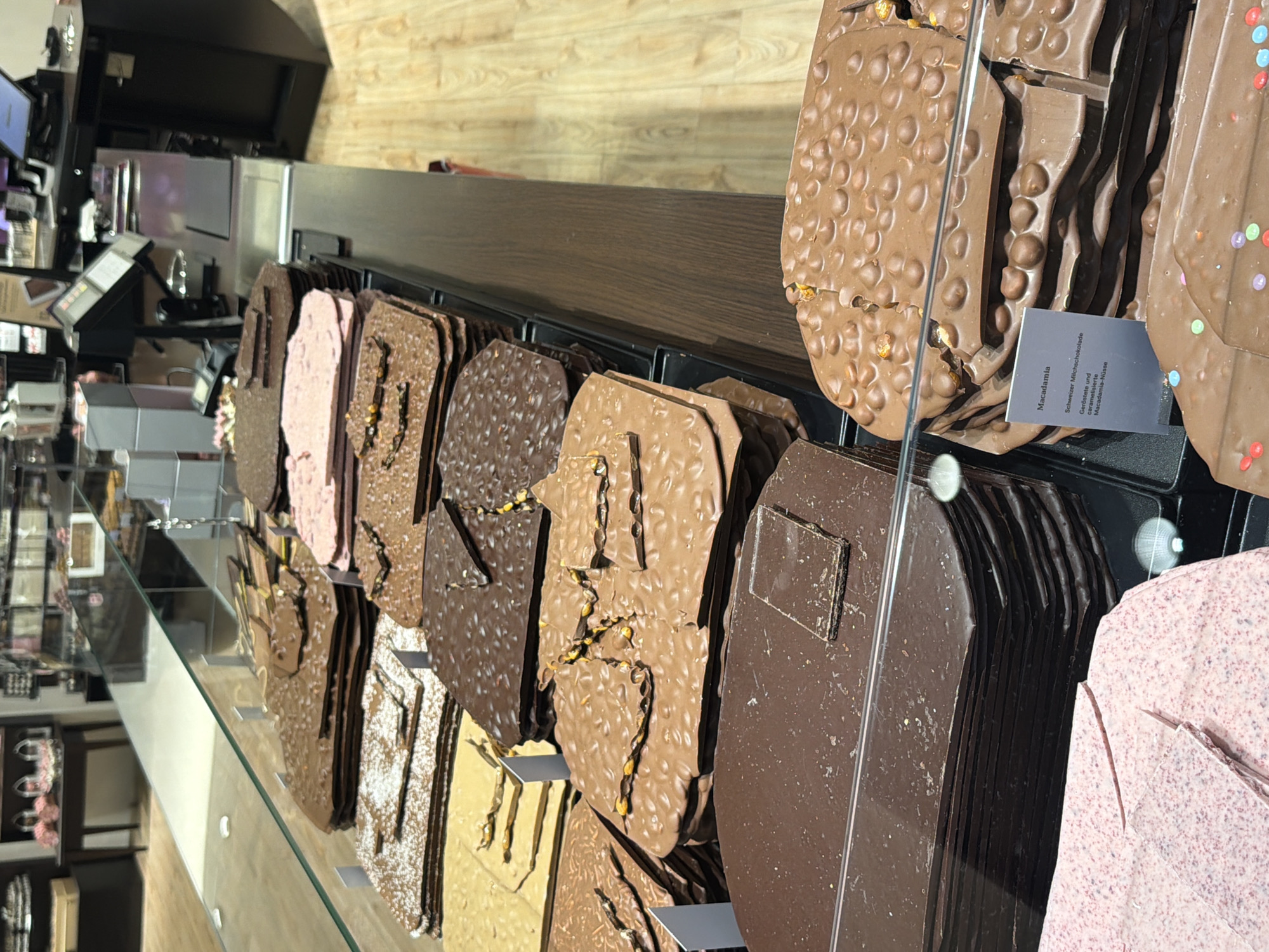 The image depicts a display case filled with various types of chocolate slabs, likely in a confectionery or chocolate shop. The chocolates vary in color and texture, some featuring nuts and other toppings. A small label, partly visible, identifies one type as "Macadamia." The location is suggested to be in Spreitenbach, Switzerland. The scene is viewed through the glass of a display case, with reflections on the surface. The background includes a blurred view of a store interior.