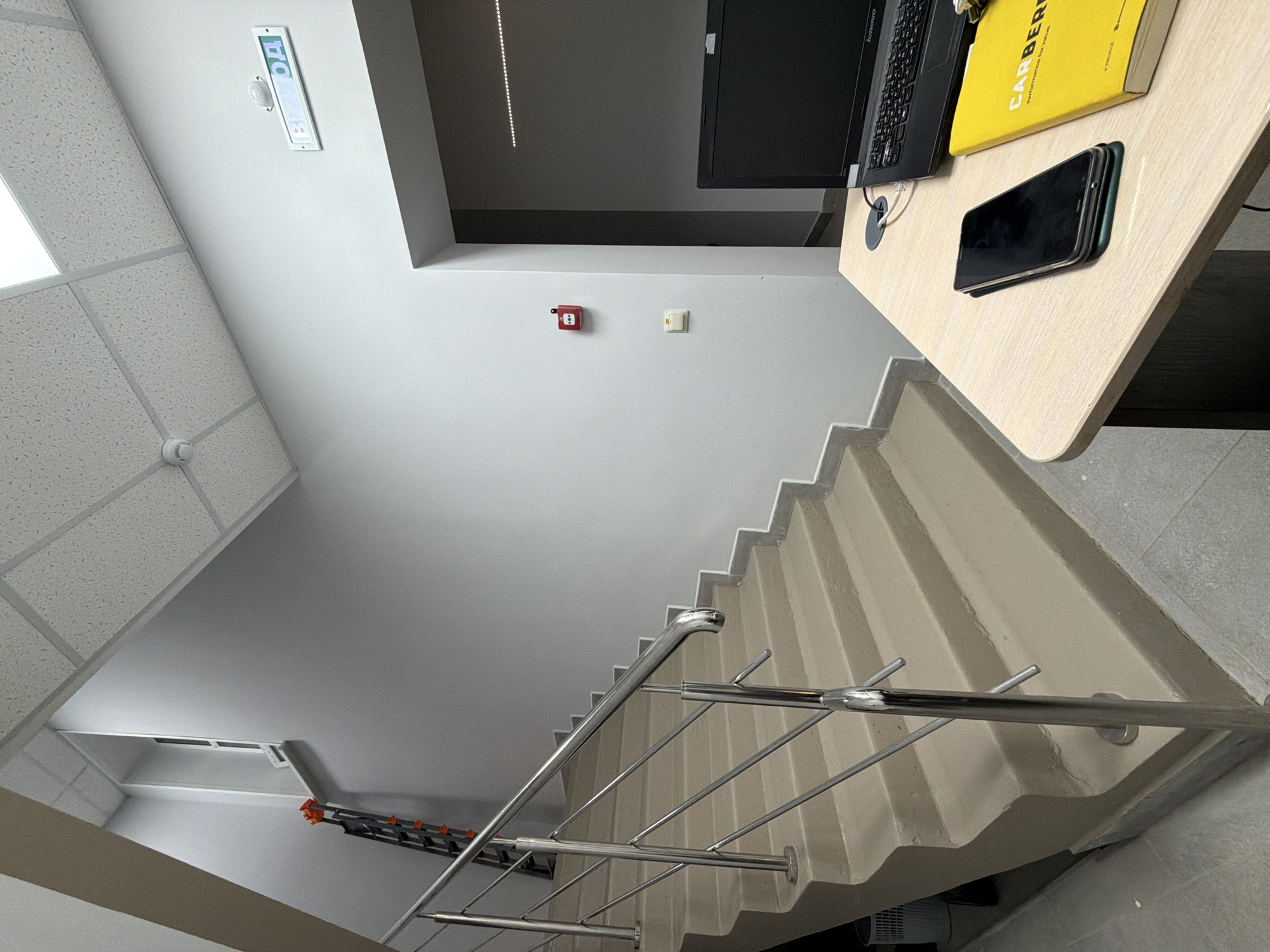 An eye-level shot inside a building, showcasing a staircase with a silver railing that leads upwards. The staircase is light beige, contrasting with the gray wall beside it. In the upper right corner, part of a table is visible with a laptop, a yellow book, and what appears to be a mobile phone on top. The upper left corner reveals a white tiled ceiling with embedded lighting fixtures. A rectangular sign with green lettering hangs on the wall, possibly indicating an exit or direction. Towards the middle of the wall, there are a red fire alarm pull station and an adjacent white electrical outlet. In the background, toward the lower left, there is a glimpse of a white storage area with an orange and black folding ladder. The specific location in Sarapul, Russia, cannot be determined from the image.