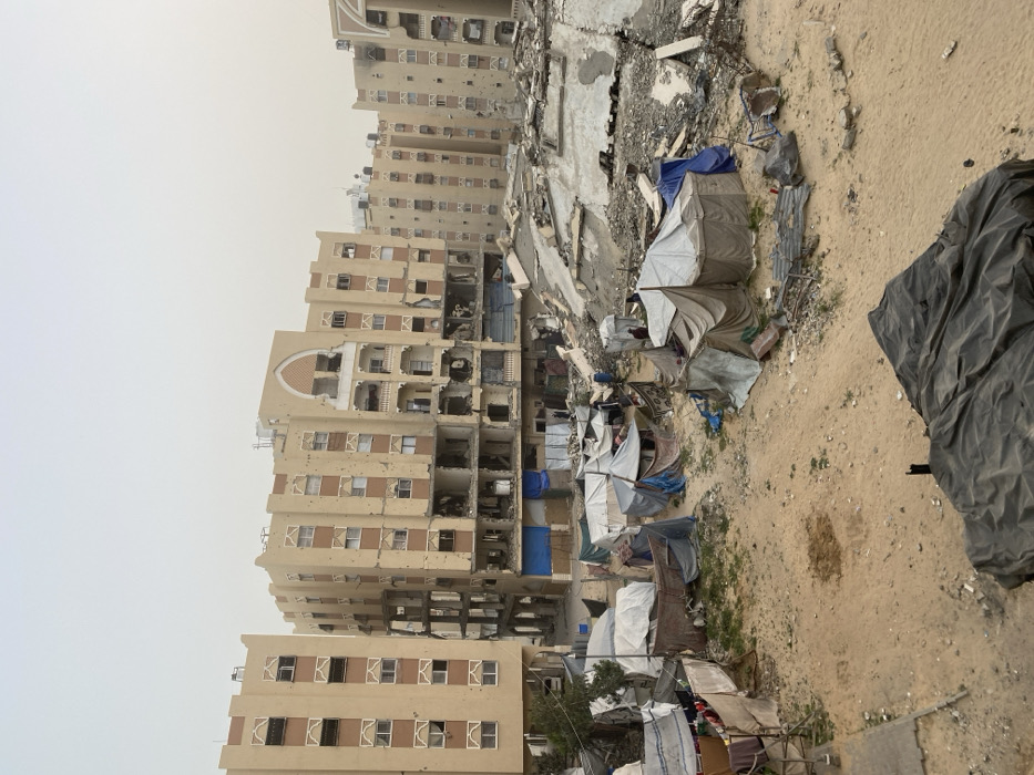 The image portrays a devastated urban environment in Khan Yunis, Palestinian Territory, characterized by extensive structural damage and makeshift living conditions. In the background, several multi-story residential buildings, constructed of light brown concrete, exhibit severe destruction. The central and right-hand buildings have large sections of their facades collapsed or blown out, exposing internal structures, and are visibly pockmarked with numerous impact holes. A substantial pile of concrete rubble and debris lies at the base of these damaged buildings. In the midground, an informal encampment of approximately 15-20 makeshift tents is set up on a sandy, unpaved area with sparse vegetation. These tents are constructed from various materials, including tarpaulins (white, grey, blue) and other fabrics; laundry is visible hanging outside some structures. Several indistinct individuals are present among the tents. The foreground consists of sandy ground with a large, dark tarp or bundle. The sky is hazy or overcast.