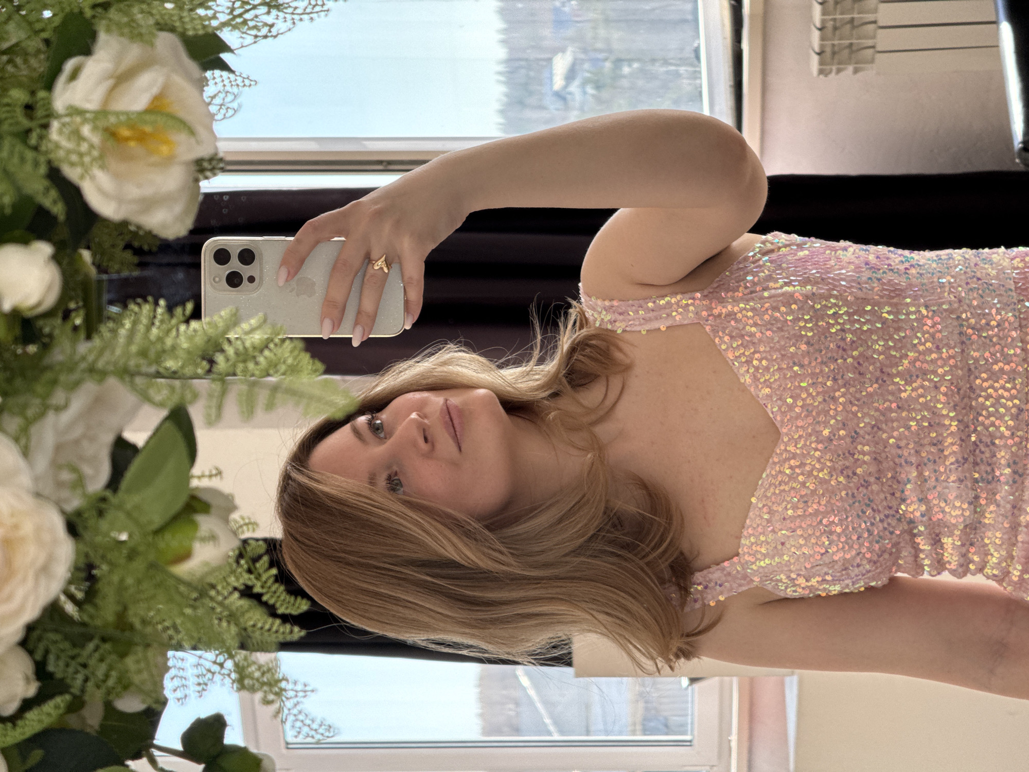A woman is taking a mirror selfie indoors. She is holding an iPhone in her left hand, with the phone partially obscuring her face. She has blonde hair and is wearing a pink sequined dress. A bouquet of white flowers is situated on the left side of the frame, partially obscuring the view.
The reflection in the mirror shows a window with a view of a city. It is likely a city in Russia based on the style of architecture visible in the background and the source’s prompt, possibly the city of Chaikovsky, Russia.
The room appears to be well-lit, with natural light coming from the window. A radiator is visible on the top right side of the image.