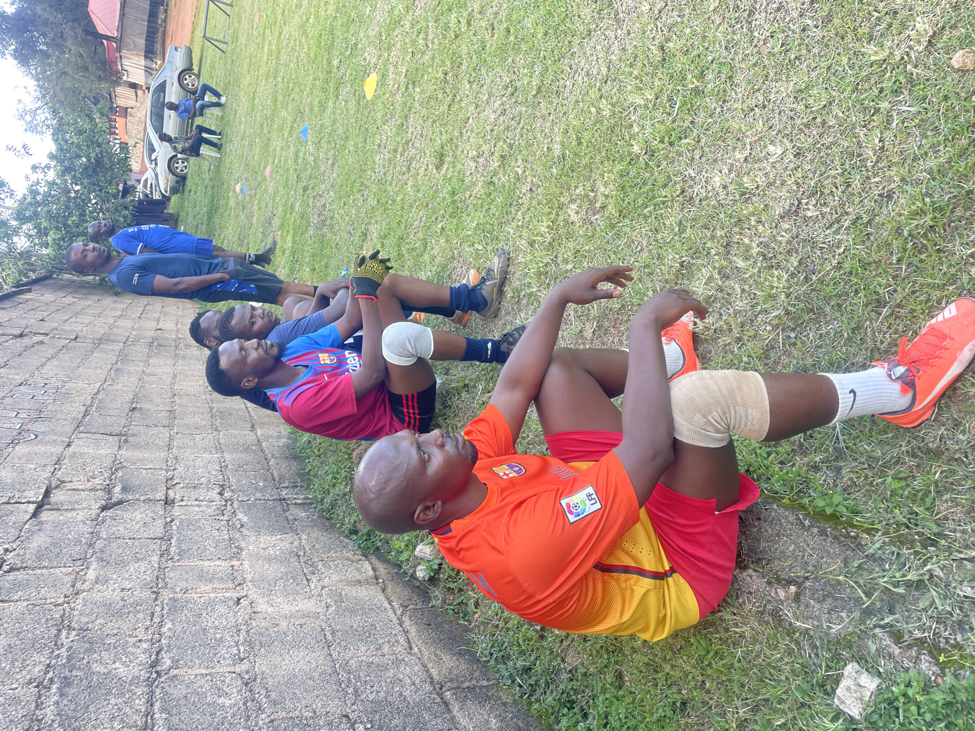 A group of men are seated on the grass next to a grey brick wall, with some standing behind them. The men are wearing athletic attire, including jerseys, shorts, and athletic shoes. Some are wearing knee pads. The environment appears to be an outdoor sports field, with grass covering the ground and trees and buildings visible in the background. Cones are scattered on the grass, suggesting a sports training session or game. The scene is illuminated by natural daylight. The man in the foreground, wearing an orange and yellow jersey, is bald and looking to the right. Another man in a blue and red jersey is seated to his left, and another in a blue jersey is standing behind them. Two men are seated further in the background, one in a blue shirt and the other in a grey shirt. The presence of athletic wear, cones, and the overall setting indicates a sporting context, likely a football or soccer practice or match. The architecture in the background, including a red-roofed building, suggests an urban or semi-urban environment. The general scene is one of rest or observation during a sporting activity. The details of the brick wall and the surrounding vegetation suggest a potentially warm climate. Without additional context, it is difficult to definitively pinpoint the exact location within Jos, Nigeria, but the presence of athletic activities and the visible urban elements are consistent with areas found in Nigerian cities.