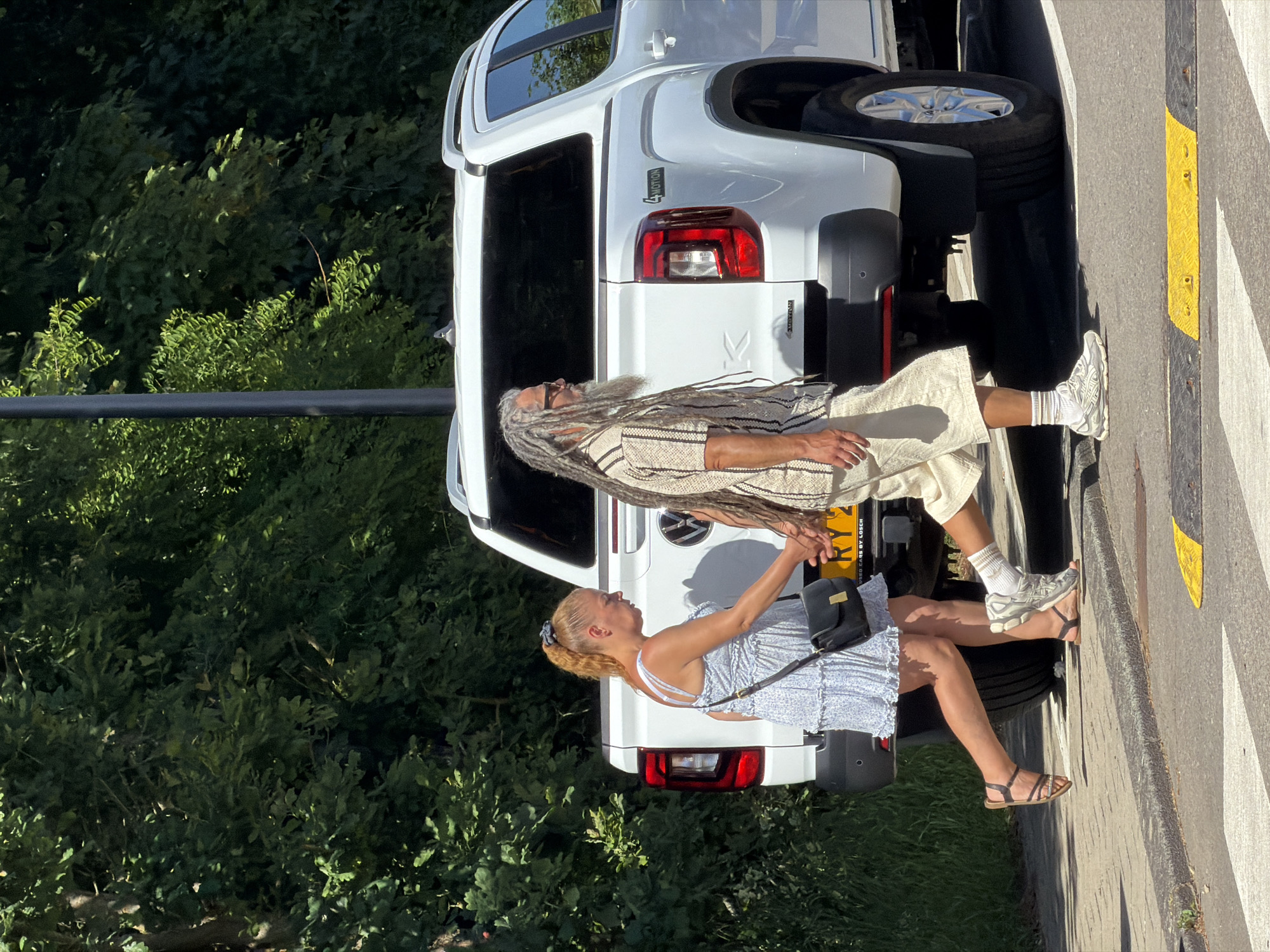 Two people are walking along a curb, next to a white Volkswagen Amarok vehicle, license plate RY2. The woman is on the side closest to the vehicle, wearing a blue and white dress, dark sandals, and a black purse. The man is walking slightly ahead of her, wearing light-colored clothing, white socks, white sneakers, and has long dreadlocks. Behind them, there is a lush green area. The curb is marked with yellow and black stripes. The location is Fentange, Luxembourg, as inscribed on the license plate.