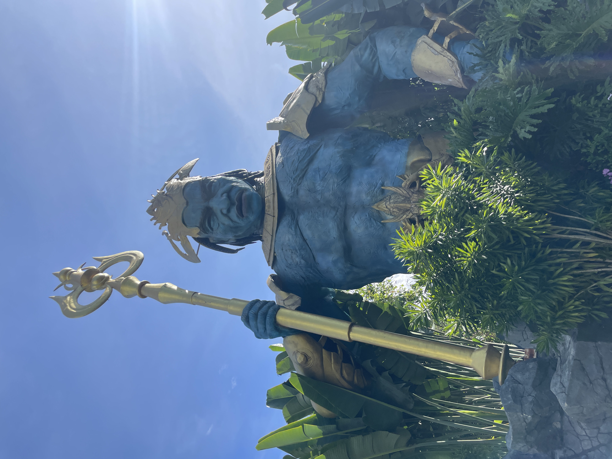 A low-angle, eye-level shot captures a colossal blue statue of a mythical warrior, likely of oceanic or aquatic origin, holding a golden trident. The statue is adorned with ornate gold armor on its shoulders and chest. It stands amidst lush green foliage and dark, textured rocks, possibly in a garden or themed attraction. The sky above is a clear blue with the sun creating a lens flare effect on the right side. The statue's facial features are stern and imposing, with dark, braided hair and a golden headdress. The image provides a detailed, realistic portrayal of this impressive monument.