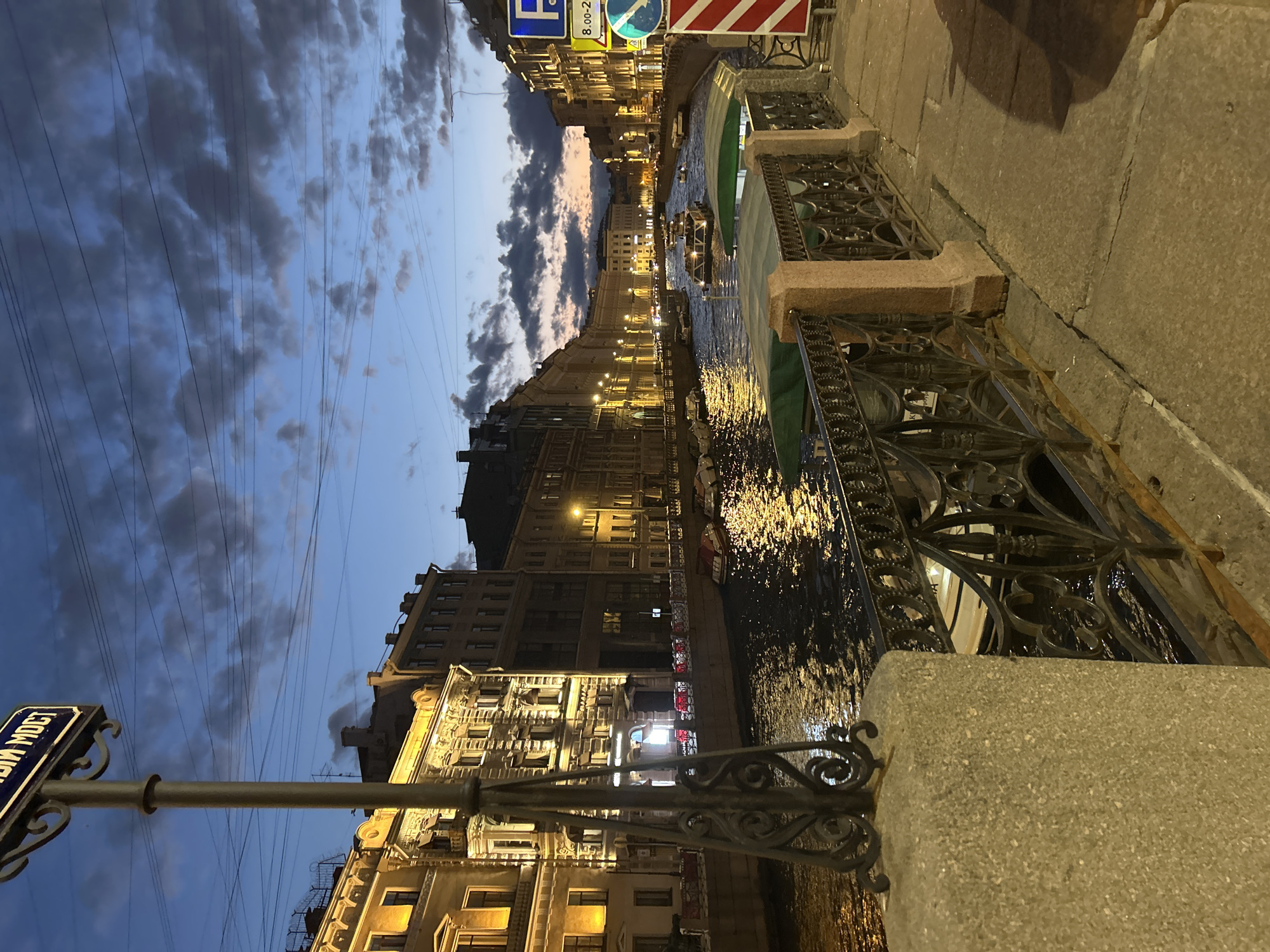 A canal-side view of a city at dusk shows illuminated buildings along the water. The sky is a deep blue with scattered clouds, and the water reflects the lights from the buildings and several boats. Ornate metal railings and stone balustrades line the waterfront promenade. A street signpost with Cyrillic lettering is visible in the foreground on the left, and traffic signs, including a blue parking sign, are present on the right. The overall scene depicts an urban environment at twilight, likely a European city.