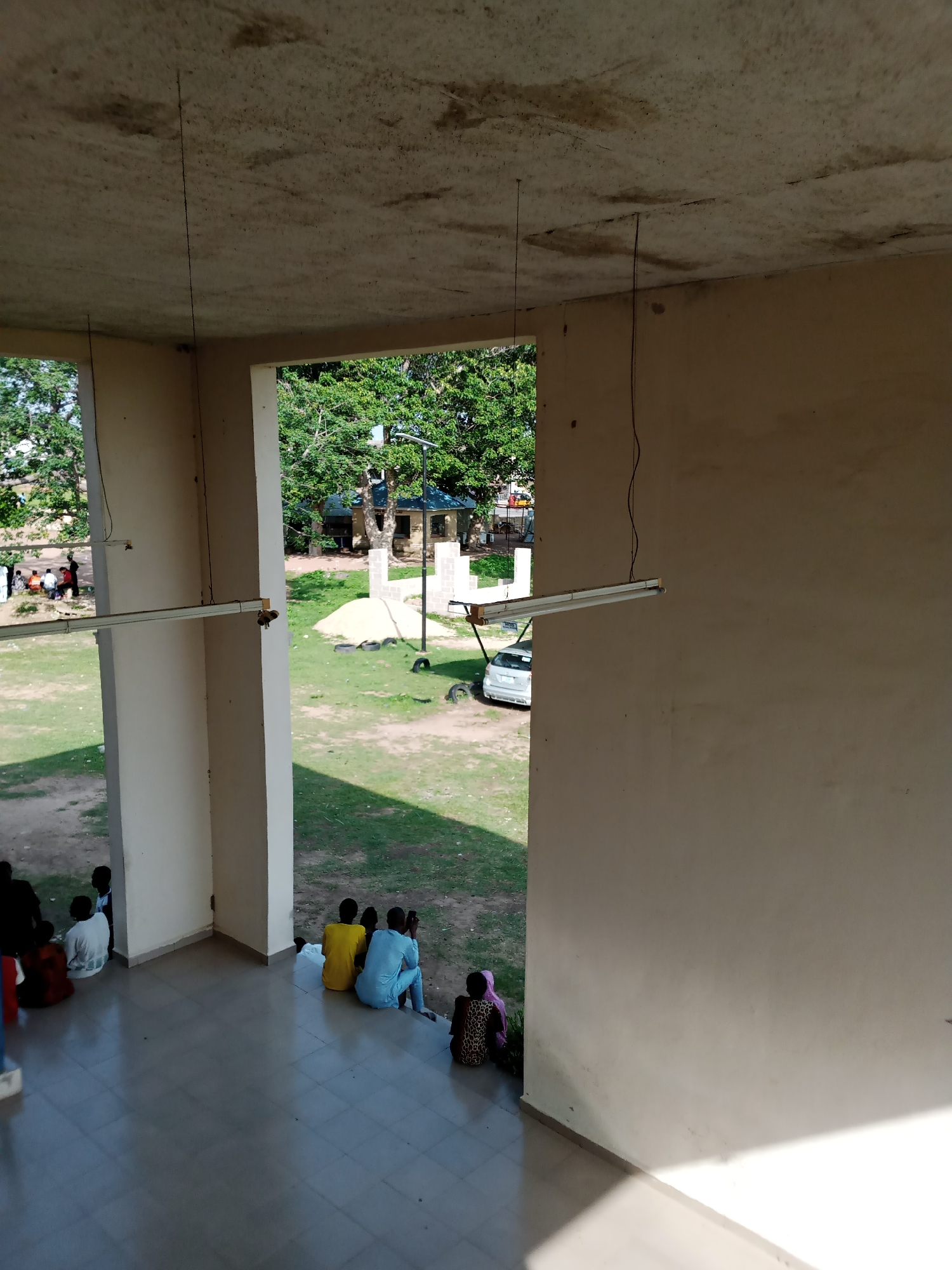 The image depicts an outdoor scene in Jalingo, Nigeria, focusing on a partially constructed or open-air building. In the foreground, a tiled floor leads to a large, rectangular opening in the building, revealing an outdoor area. Several people are seated on the steps and floor of the building's entrance, facing outward. On the exterior, a grassy area is visible, with scattered sand, some black tires, and a white car parked. Further in the background, trees and a small structure with a green roof are present. The building itself has a concrete ceiling with exposed wiring and fluorescent lights attached. The overall impression is one of a public or communal space, possibly undergoing renovation or in a state of disuse, with people gathered outside.