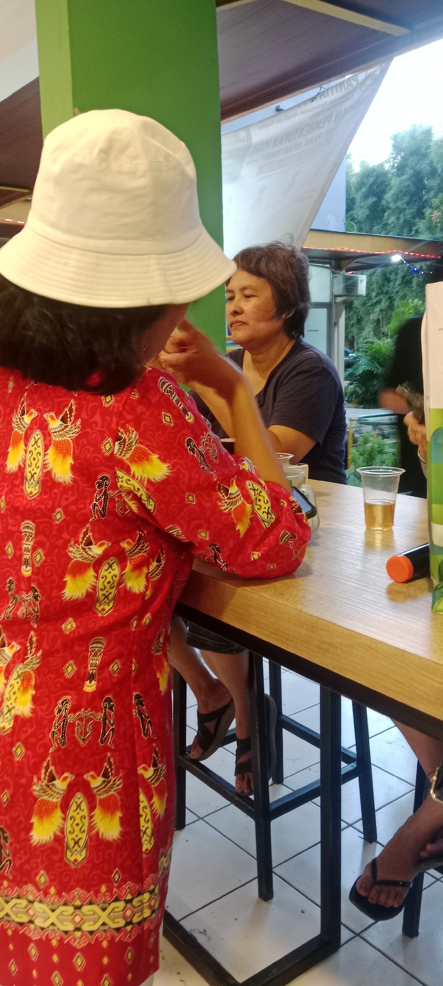 The image depicts an indoor seating area, likely a cafe or restaurant, with a natural light setting. In the foreground, a person wearing a red and yellow patterned dress and a white bucket hat is seated at a wooden table. Behind them, another individual with short, dark hair and a dark blue t-shirt is facing away from the camera, looking to their left. A plastic cup with a yellow beverage and a black cylinder with an orange tip are visible on the table. The background shows a green wall, a white banner with text, trees outside, and more seating. The flooring is white tile. The image appears to be taken in East Jakarta, Indonesia.