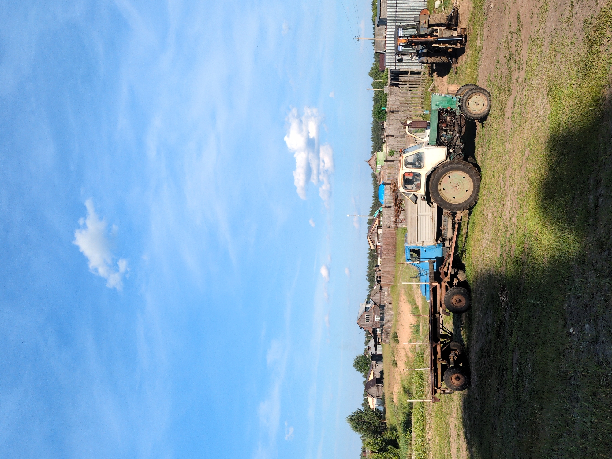 This is a vertical, eye-level, outdoor, daytime, full shot of a rural scene in Molochonoy Fermy, Russia. The sky is bright blue with scattered white clouds. In the foreground, a large shadow is cast across a grassy field.
In the middle ground, a blue truck with a long trailer is parked on the grass. To the right of the truck, a green and white tractor with a person in the driver's seat is also parked. Further to the right, another tractor with a front loader is visible. Behind the vehicles, a wooden fence separates the yard from a collection of rustic wooden houses and buildings. Trees and more wooden fences are visible in the background, suggesting a rural village. The overall impression is one of a working farm or agricultural area.