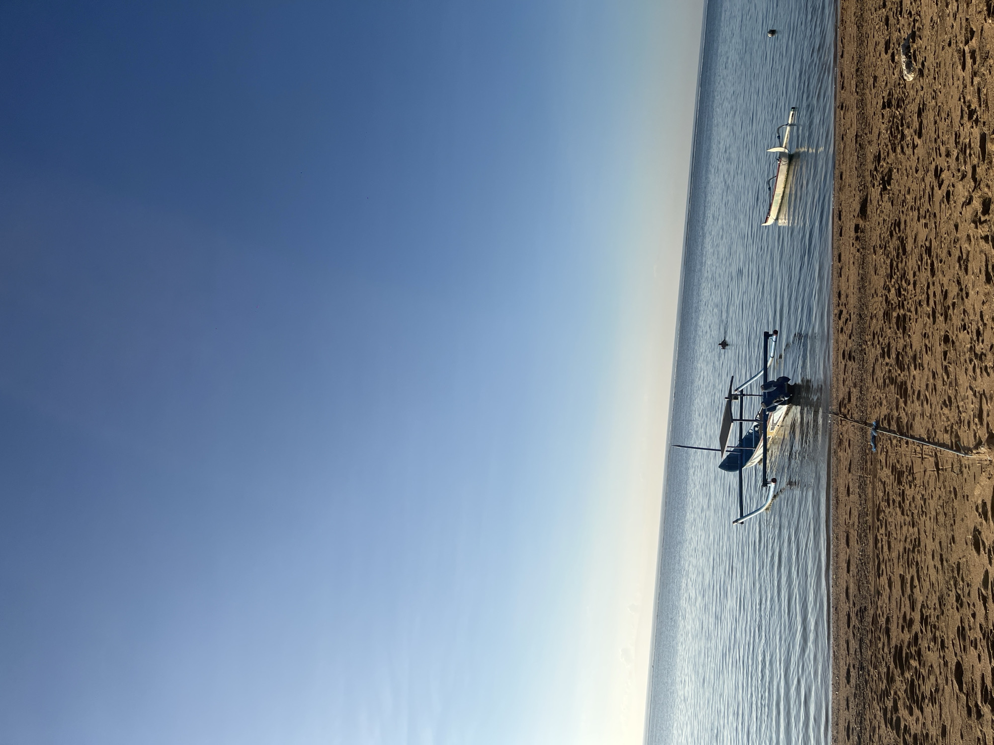 A tranquil scene unfolds on a beach in Denpasar, Indonesia, under a clear blue sky. The foreground is dominated by a sandy beach, marked with subtle footprints and a fishing rod set up near the water's edge, suggesting a leisurely morning. The calm, shallow water gently laps at the shore, reflecting the soft light of the sky.
Two traditional Indonesian outrigger boats are visible. The larger boat, predominantly blue with white accents, is anchored closer to the shore. It features a simple canopy structure and an outboard motor. The second, smaller boat, white with red trim, floats further out, appearing serene and empty. In the distance, a lone swimmer can be seen in the water, adding a subtle human element to the otherwise quiet seascape. The horizon is a soft blend of sea and sky, with distant landmass or clouds faintly visible on the horizon. The overall atmosphere is one of peaceful serenity, capturing a typical morning on Bali's coast.