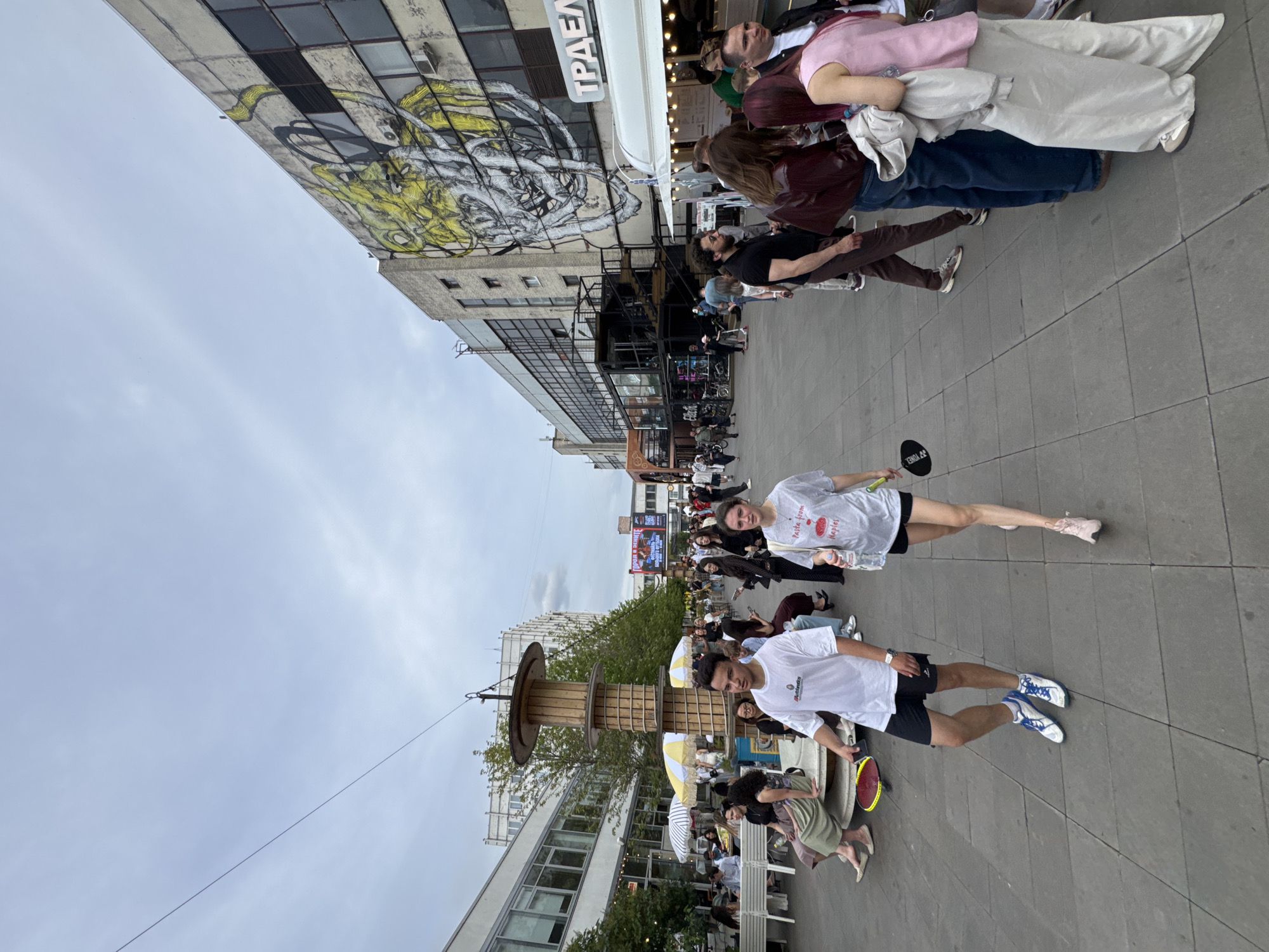 The image depicts an outdoor scene in Saint Petersburg, Russia, on an overcast day. A group of people are gathered, suggesting a social event or a casual outing. The setting appears to be an urban area with modern architecture, including a building with graffiti on its facade and another with many windows. Some people are walking, while others are seated or standing around.
In the foreground, a man in a white t-shirt and black shorts is walking, wearing athletic shoes. Beside him, a woman in a white t-shirt is also walking, her legs visible. She is holding a black object with a logo that is difficult to discern. Further back, several other individuals are present, some engaged in conversation, others observing their surroundings.
A notable object in the scene is a large, wooden reel, possibly used for industrial purposes, which has been repurposed as a decorative element or a stage. Two small, yellow and white striped umbrellas are placed near the reel, adding a touch of whimsy to the urban landscape. The overall atmosphere seems relaxed and communal.
Some visible text includes the word "ТР AD EM" on a white awning above a building, likely part of a shop or restaurant sign. The woman in the white t-shirt has text on her shirt that reads "Taste from Naples." The man in the foreground has a logo on his t-shirt that is not entirely clear but appears to be a brand name. The weather is cloudy, indicating a typical Saint Petersburg sky.