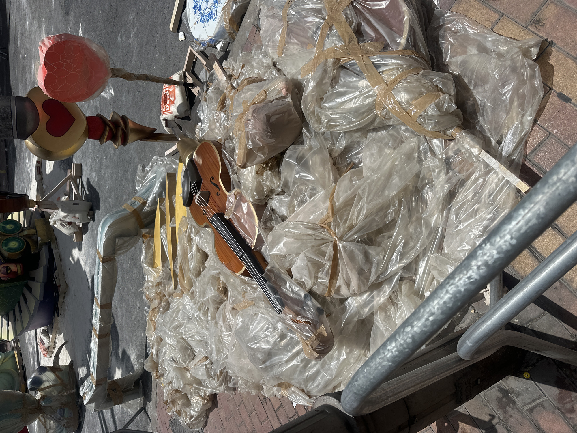 This media file depicts a close-up, overhead view of various objects, primarily decorative elements, lying on a paved surface under bright daylight. The scene appears to be an outdoor setting, possibly a workshop or storage area, with a mix of partially constructed or disassembled items.
The prominent subjects include a golden scepter-like object topped with a large, stylized red heart shape wrapped in translucent pink material. Next to it, a violin is visible, its wooden body and strings clearly discernible, also partially covered in plastic. A large pile of items is covered in clear plastic sheeting, secured with tan tape, obscuring most of their details but suggesting a collection of assorted decorative pieces. Some of these wrapped objects appear to be layered, with some lighter colored material showing through the plastic. In the background, a wooden crate and a section of a blue and white patterned ceramic tile or plate can be seen. Other partially visible elements include what looks like part of a keyboard and some circular green decorative objects on the left side. Metal railings or supports are visible in the foreground and on the right side of the image.
The activity suggested is storage or preparation of these decorative items, likely for an event or display. The bright daylight indicates it is daytime, and the strong shadows suggest a sunny environment. The presence of what appear to be components of elaborate decorations, potentially for a festival given the Alicante/Alacant, Spain context, is implied. No people are visible in the frame. There is no discernible text in the image.