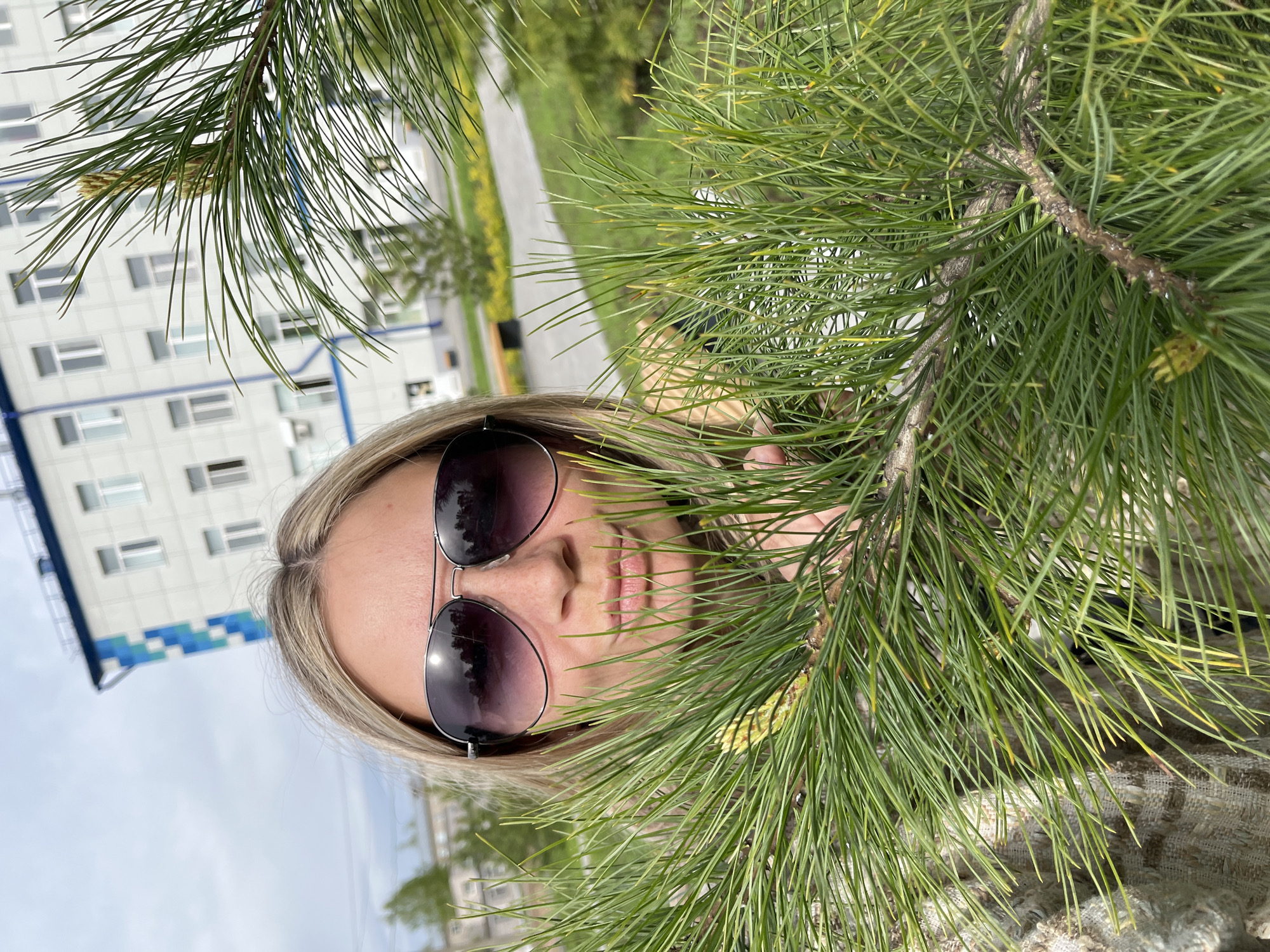 A woman with blonde hair and sunglasses partially obscured by pine tree branches is shown in an eye-level, medium shot. The branches fill the right side of the frame, with the woman's face appearing on the left, slightly behind the branches. A building with white walls and blue accents is visible in the background. In the background, there is a road with trees. The image was taken in Chaikovsky, Russia.