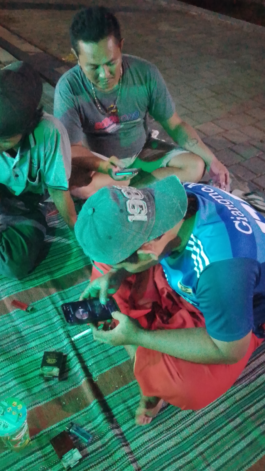 In the city of Karanglangu, Indonesia, three men are seated on a mat on the ground. The man in the foreground wears a blue shirt with white stripes and a green baseball cap, looking down at a smartphone in his hands. To his left, a man in a green shirt and dark headwear also holds a smartphone. A third man, in a grey t-shirt and a necklace, is seated further back and is also looking at his phone. Several lighters and a pack of cigarettes are scattered on the mat near them. The lighting suggests it is nighttime or a dimly lit indoor environment.