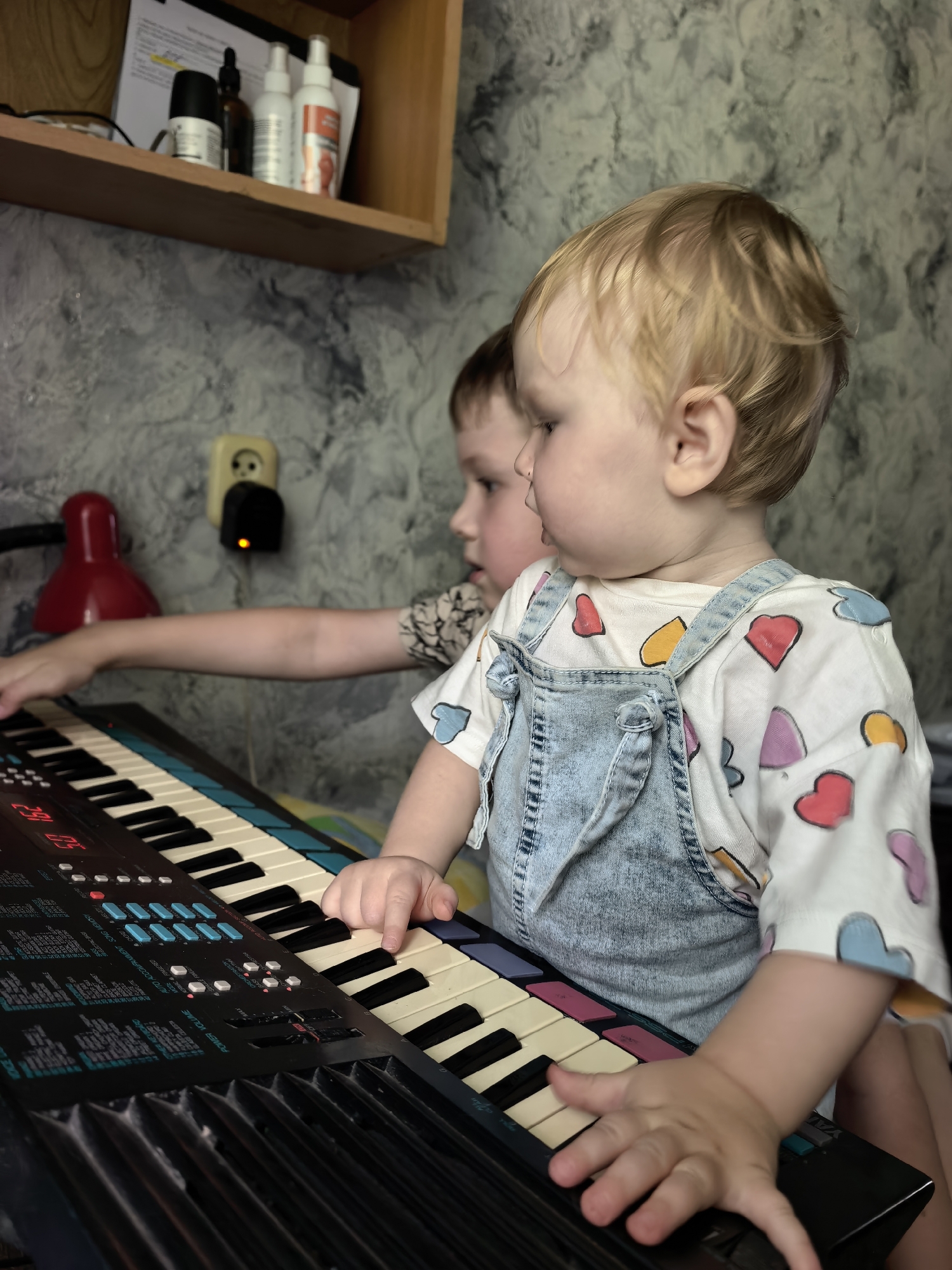 Two young children are shown interacting with a keyboard synthesizer in a room. The foreground is dominated by a toddler with blonde hair, wearing denim overalls over a white shirt decorated with colorful hearts. The toddler's hands are positioned on the keyboard, suggesting they are playing it. Behind the toddler, another child, whose face is partially obscured, is also reaching towards the keyboard. The background includes a textured wall, a wooden shelf with several bottles and papers, a red lamp, and an electrical outlet. The scene appears to be indoors, likely in a domestic setting. The specific location in Sterlitamak, Russia, cannot be determined from the visual information provided.