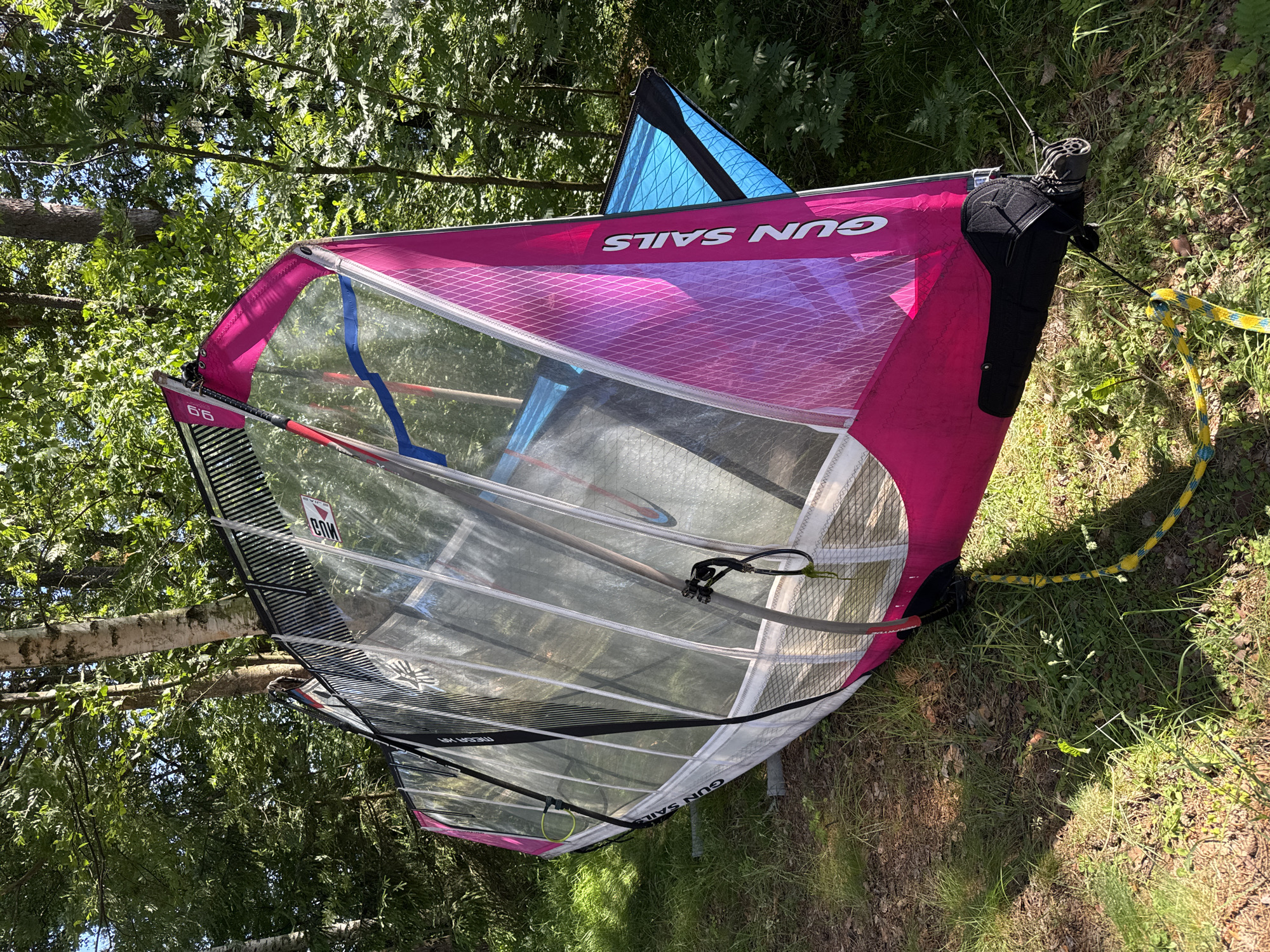 A windsurfing sail lies on the ground amidst lush green foliage and trees, suggesting an outdoor setting. The sail is predominantly pink with clear panels and features the text "GUN SAILS" printed in white. A smaller blue triangular sail is visible attached to the main sail. The scene is bathed in sunlight, indicating daytime, and the presence of grass and trees points to a natural environment, possibly a park or a campsite. The sail appears to be resting, perhaps after use or in preparation for storage. There are no people visible in the image, and the overall mood is peaceful and still. The cropped images highlight specific details of the sail, including its construction and branding, as well as the surrounding greenery. The mention of Zelenaya Roshcha, Russia, provides geographical context for the location.